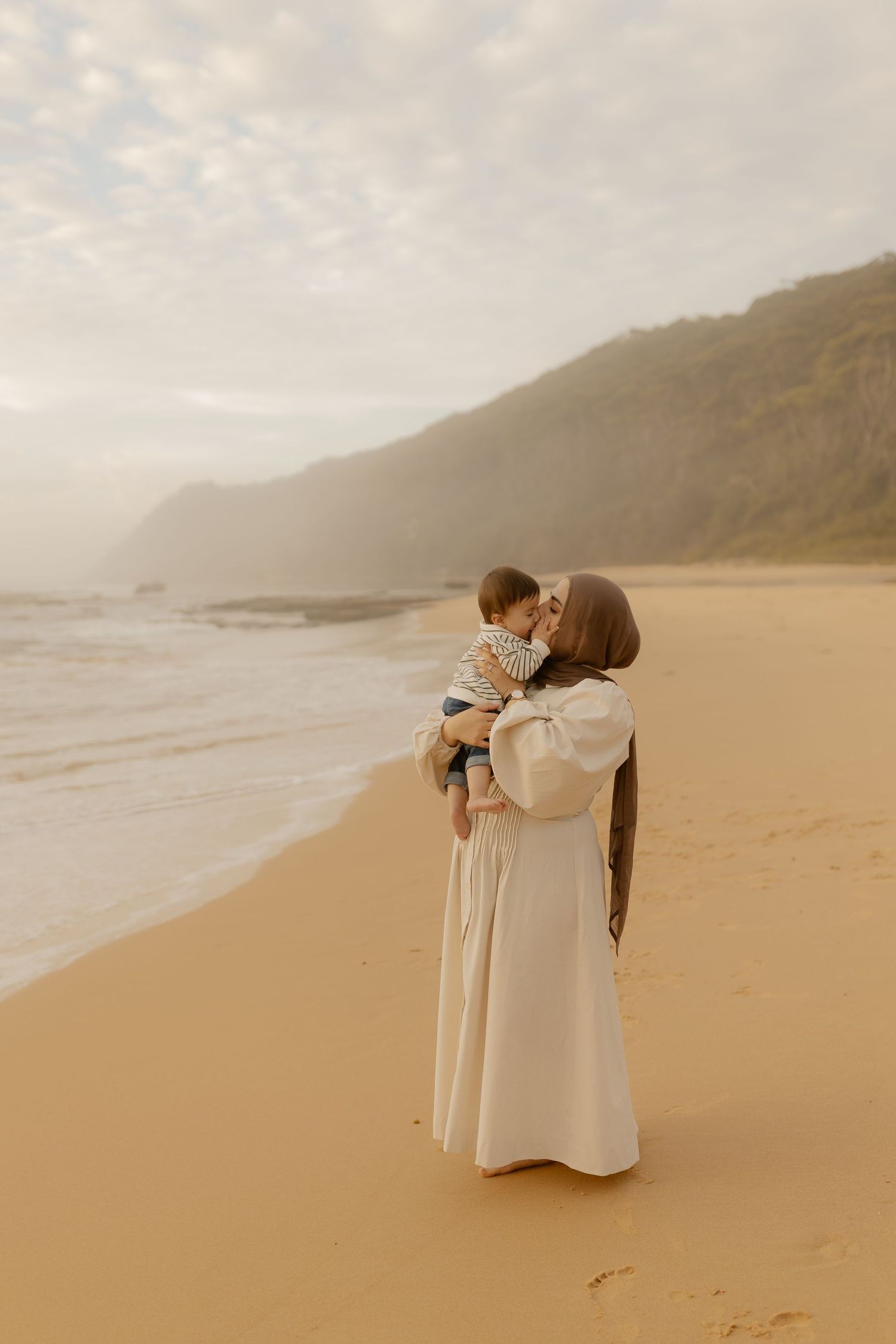 Mother in white dress holding baby on sandy beach with misty mountains and ocean waves in background.