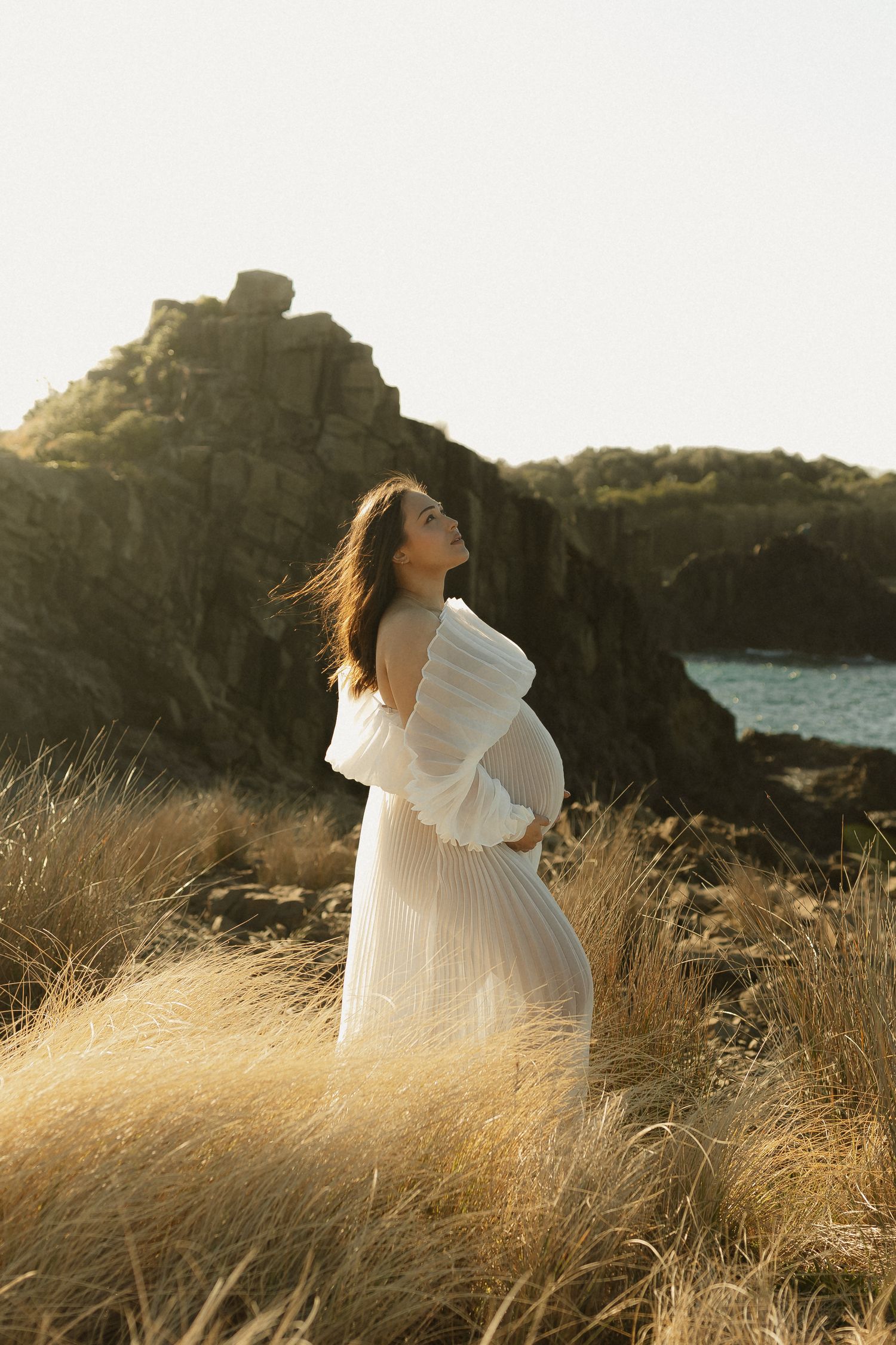 Elegant maternity photo of woman in white dress against dramatic coastal landscape.