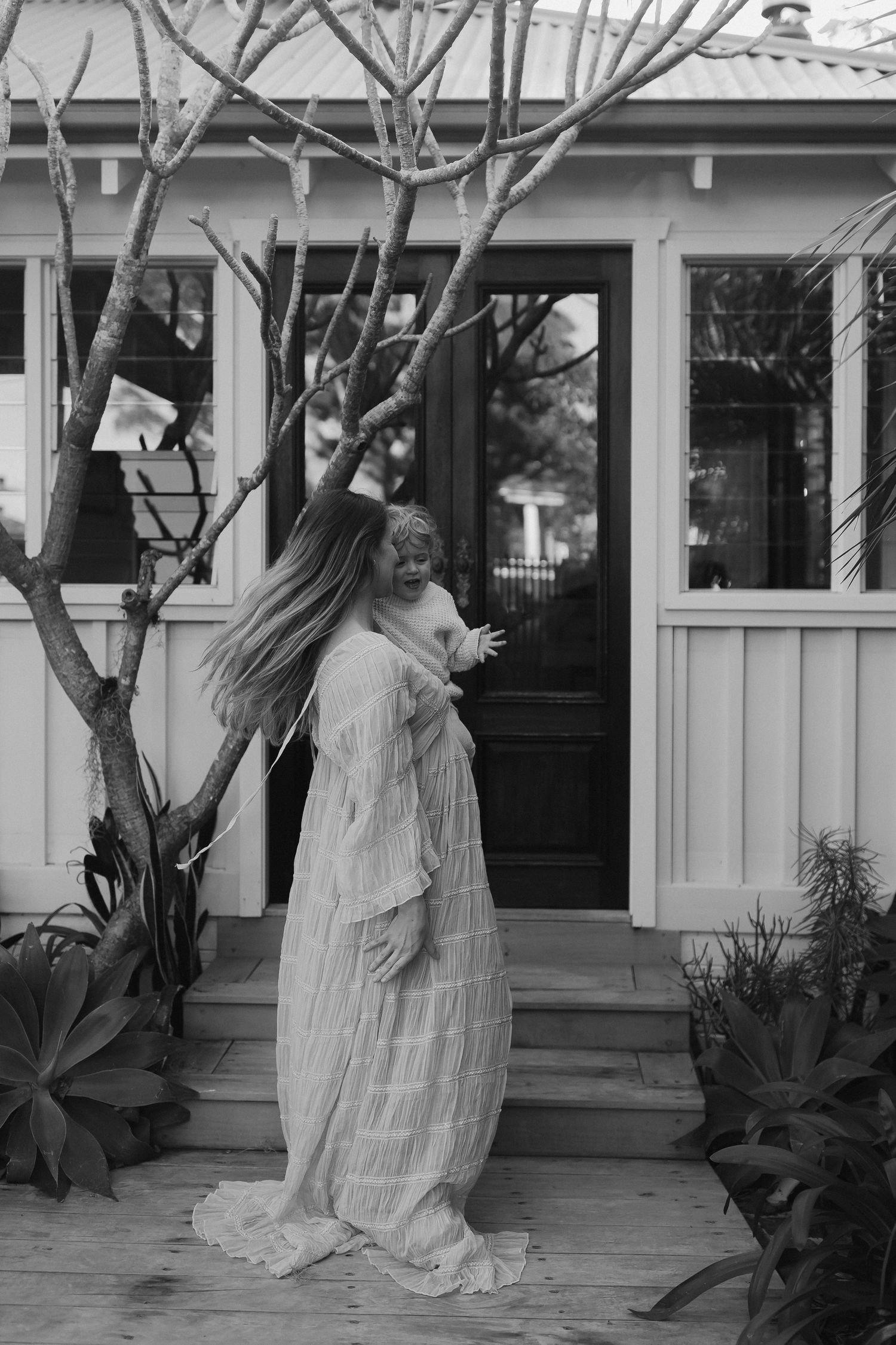 A figure in a flowing white dress moves gracefully on a porch with bare trees in black and white.