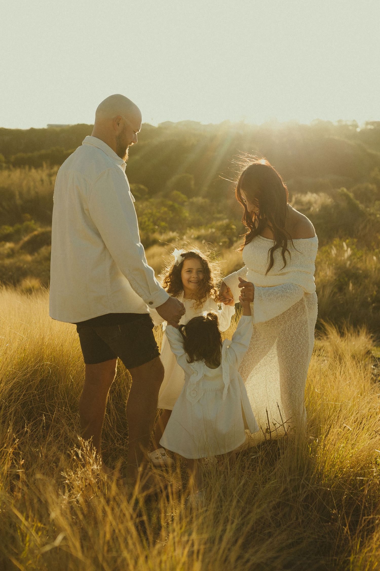 Family playing together in golden sunlit field during sunset portrait session while wearing white outfits.