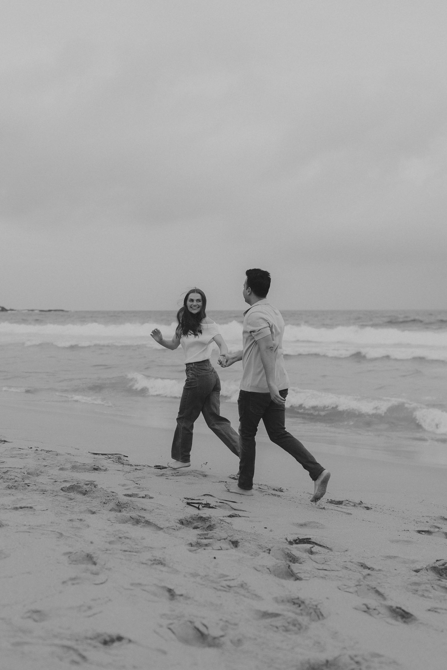 Two people playfully run and dance together along a misty beach shoreline in black and white.
