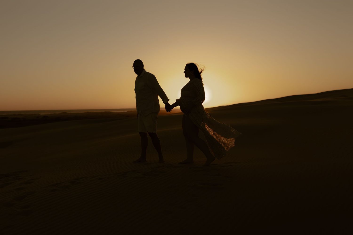 Silhouetted couple holding hands walking together against a golden sunset sky over rolling hills.