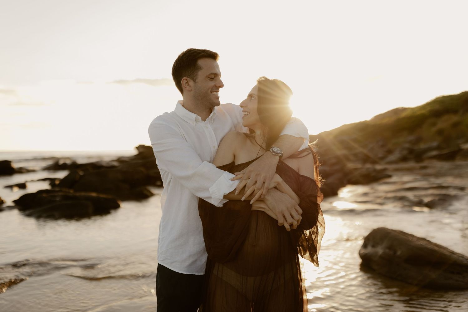 A couple shares an intimate moment on rocky beach cliffs during golden hour sunset.