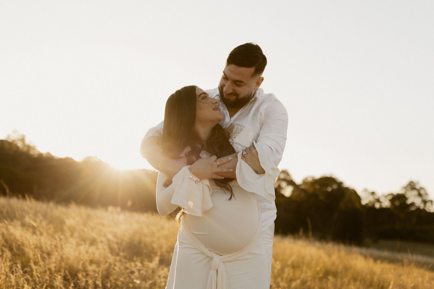 A romantic couple embraces in a sun-drenched field during golden hour creating a dreamy silhouette effect.