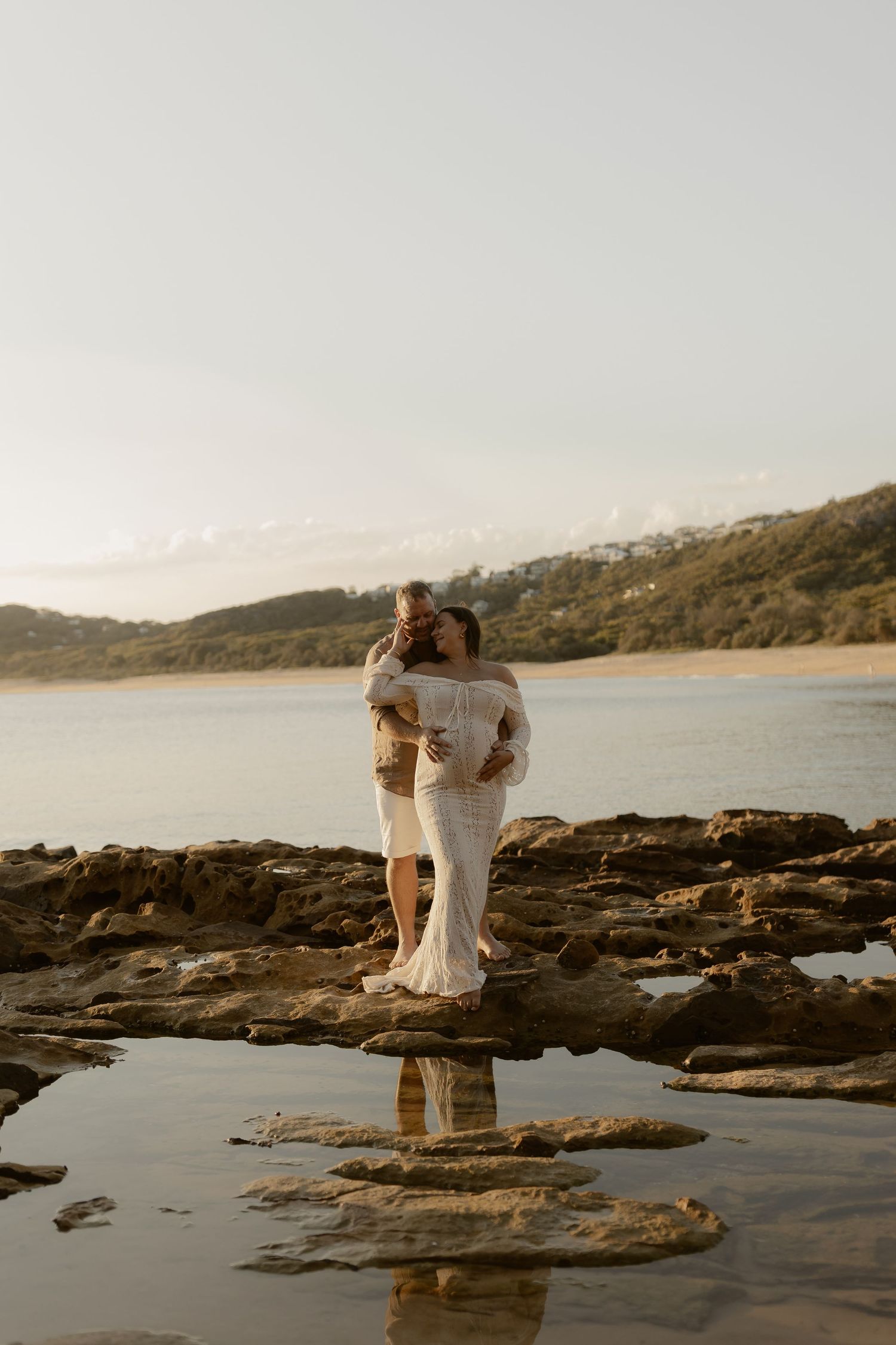 Couple embracing on rocky coastal outcrop with ocean view and dramatic sunset reflection in tide pools.