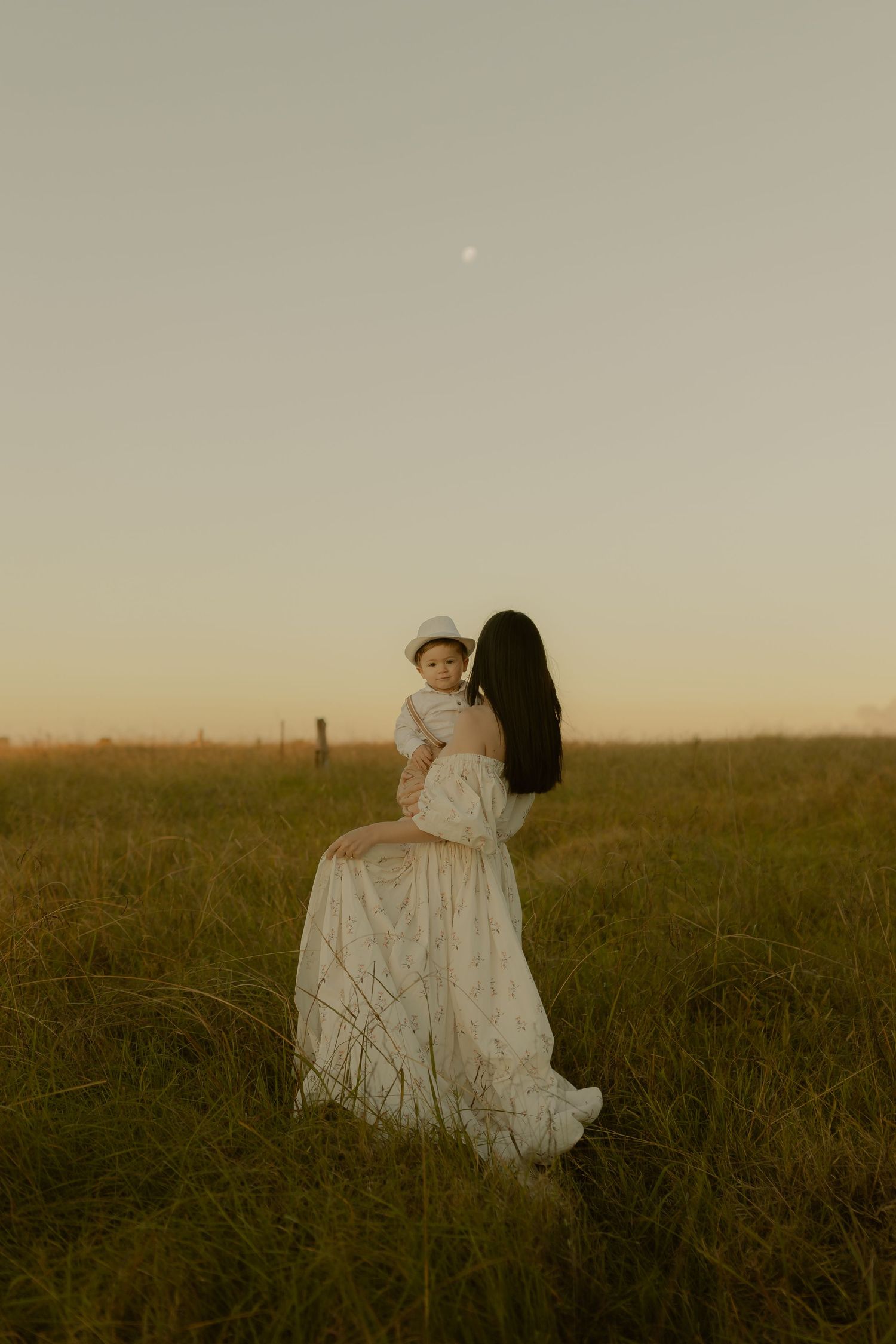 A woman in a floral dress holds a baby in a sunlit field at golden hour with the moon visible.