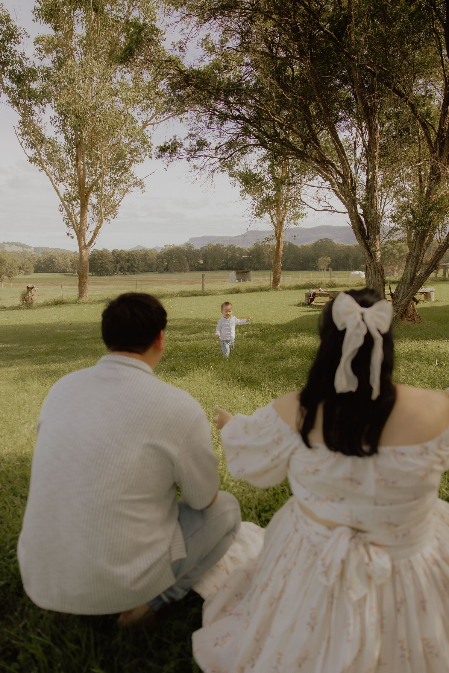 Couple sitting on grass watching toddler run across green field between trees.