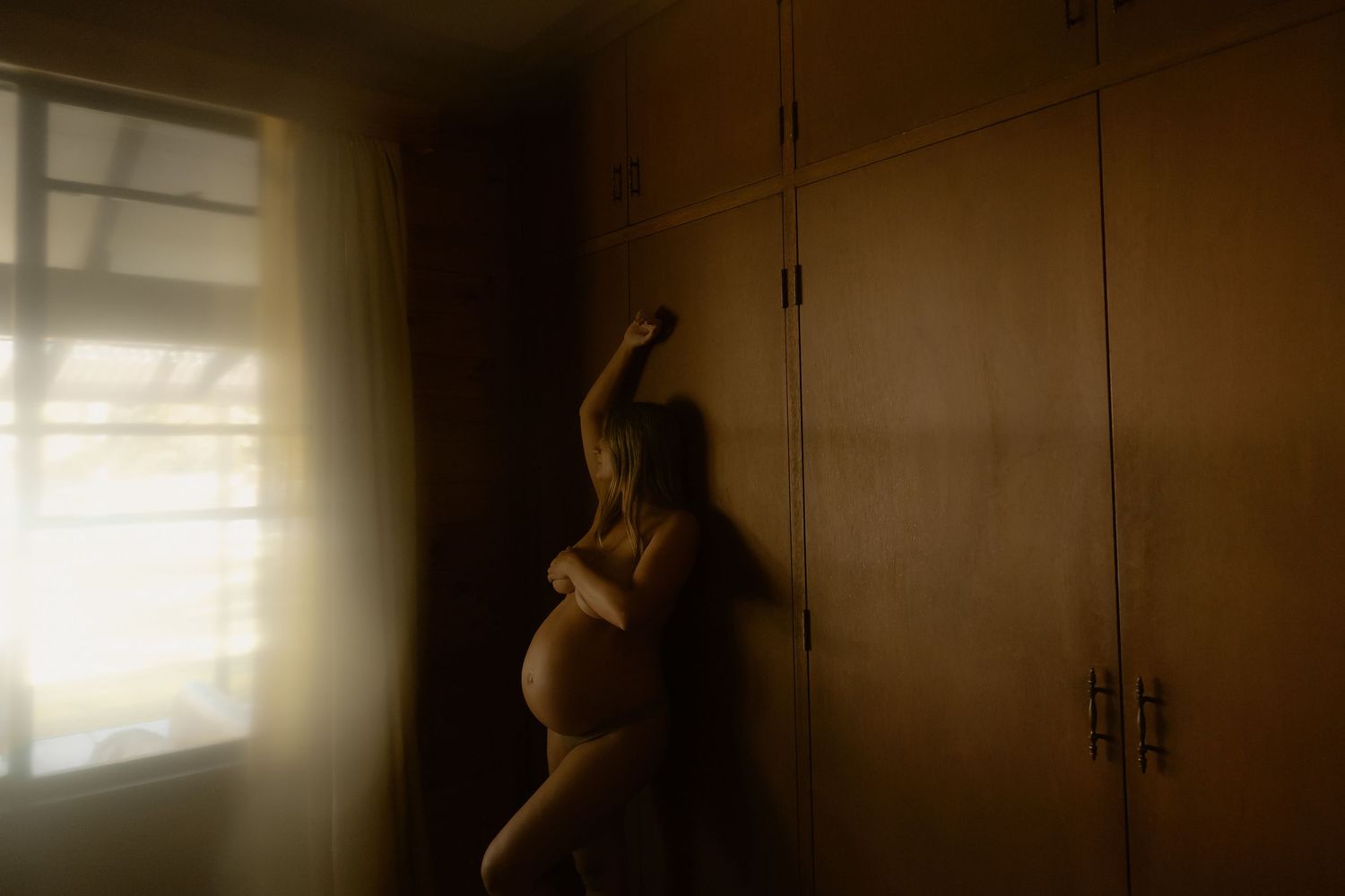 Silhouette of a nude pregnant woman leaning against wooden wardrobe doors in soft window light.