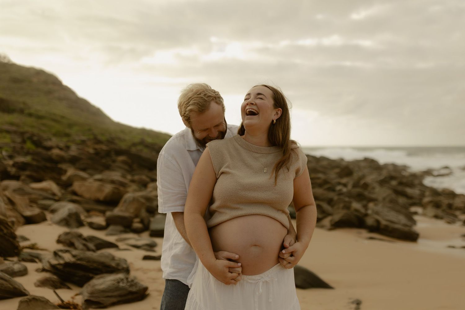 A couple shares a tender moment on a rocky beach at sunset with cliffs in the background.