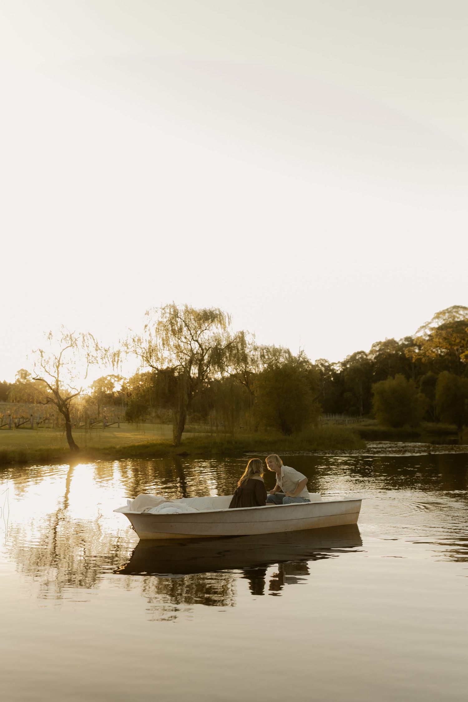 Two people sharing a romantic moment in a white boat on a serene lake with willow trees at sunset.