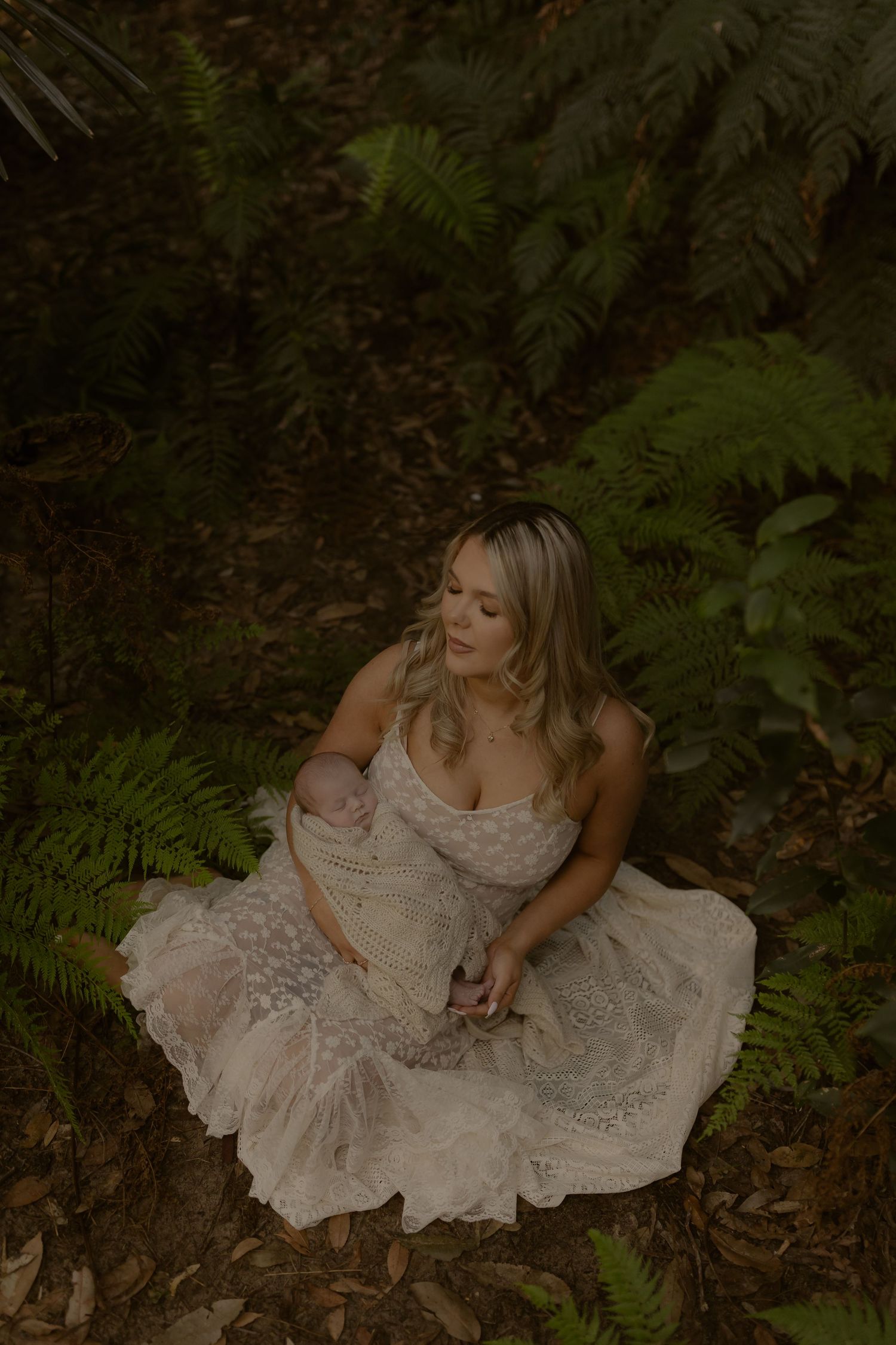 Blonde woman in elegant lace dress sits among ferns in forest, tenderly nursing her newborn baby.