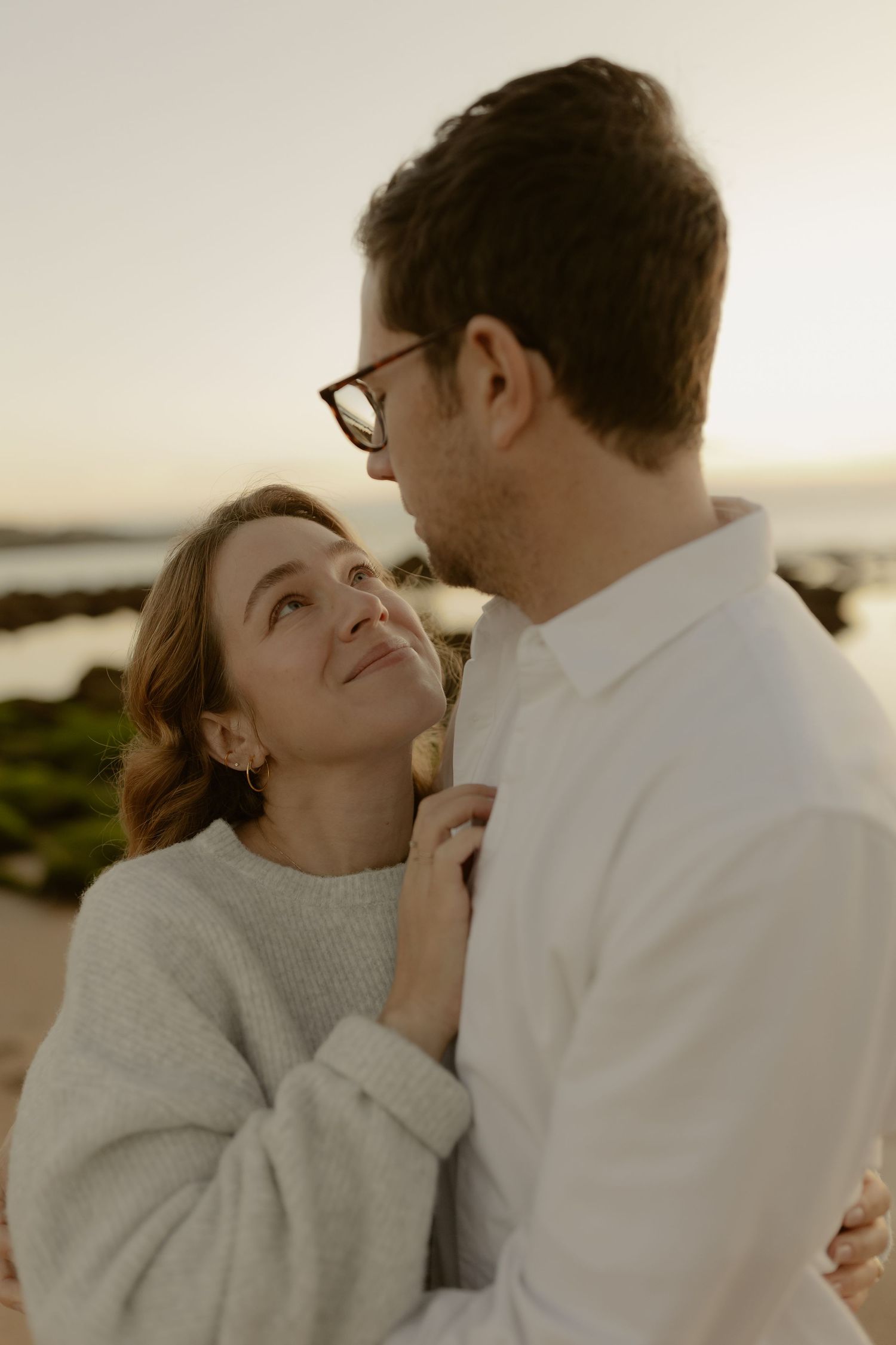 Pregnant woman in gray sweater gazing up at partner on beach during golden hour sunset.
