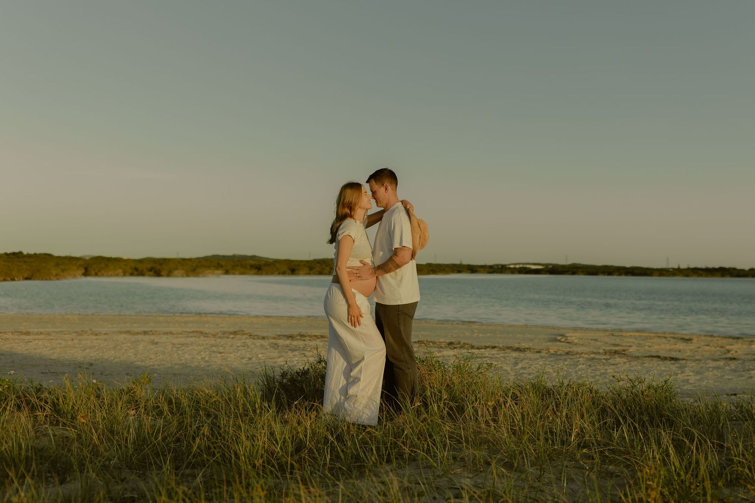 Couple embracing at golden hour on a sandy beach with calm water and green grass in the background.
