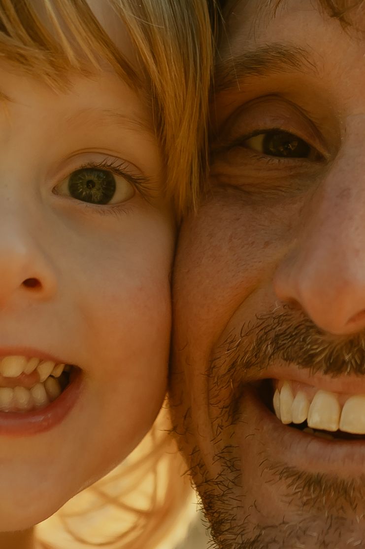 Close-up portrait of a smiling father and young child with matching green eyes and blonde hair cheek to cheek.