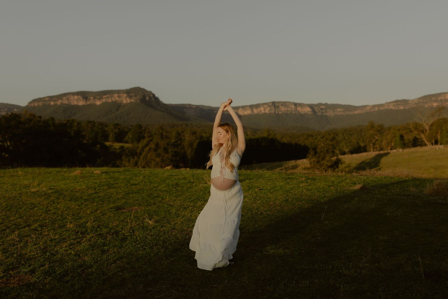 Pregnant woman in white dress dancing gracefully outdoors with mountain landscape at golden hour.