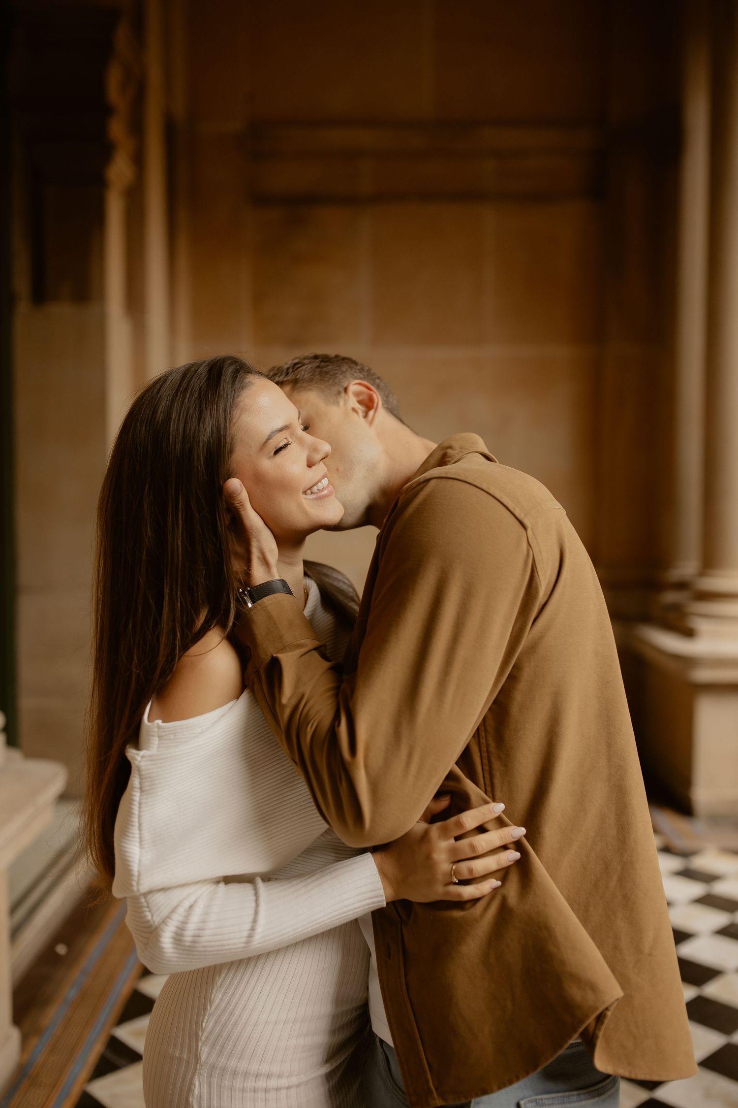 A romantic couple in matching brown and white clothing share an intimate embrace in a hallway with columns.