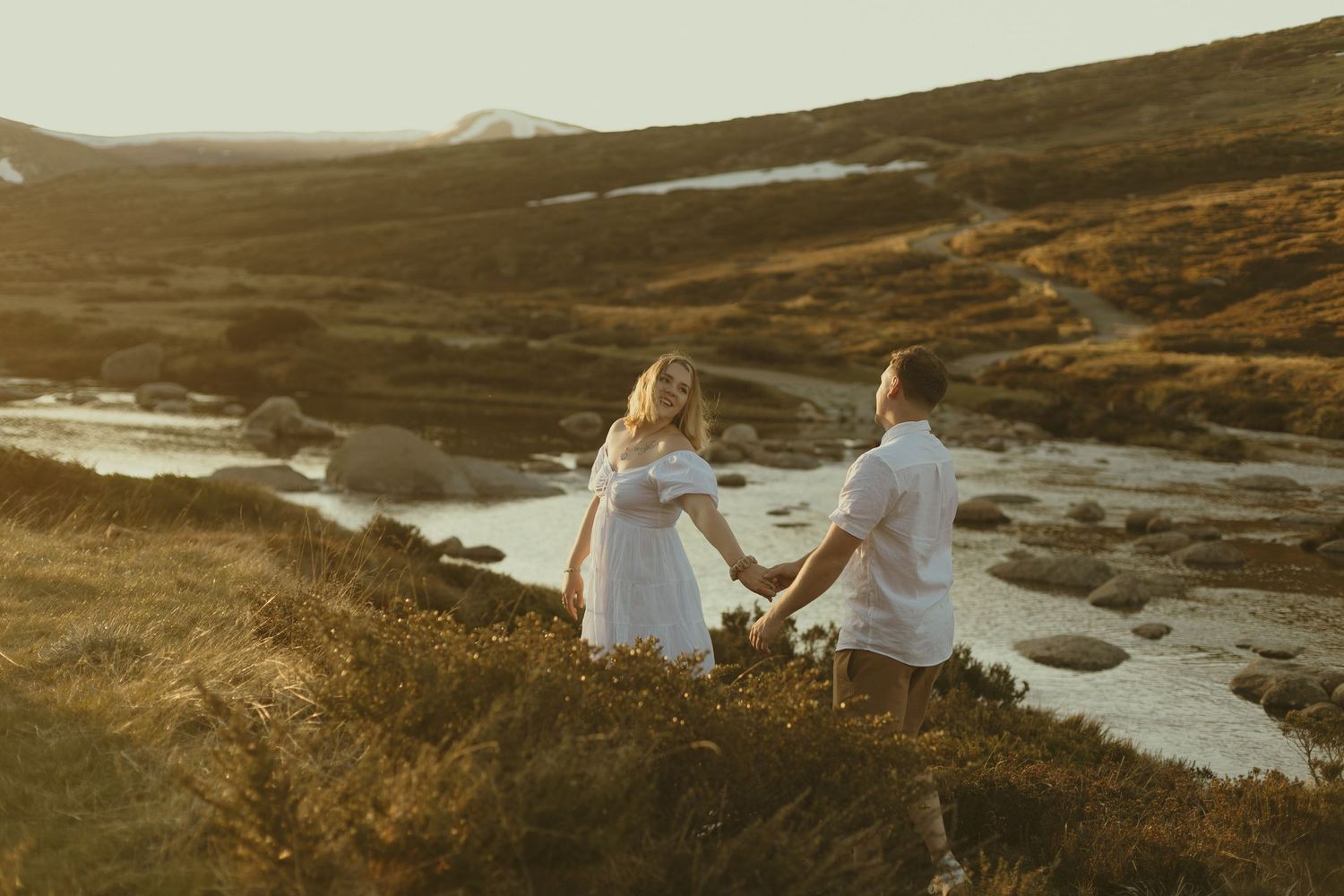 Two people in white outfits hold hands while standing on a hillside overlooking a scenic landscape with melting snow patches.