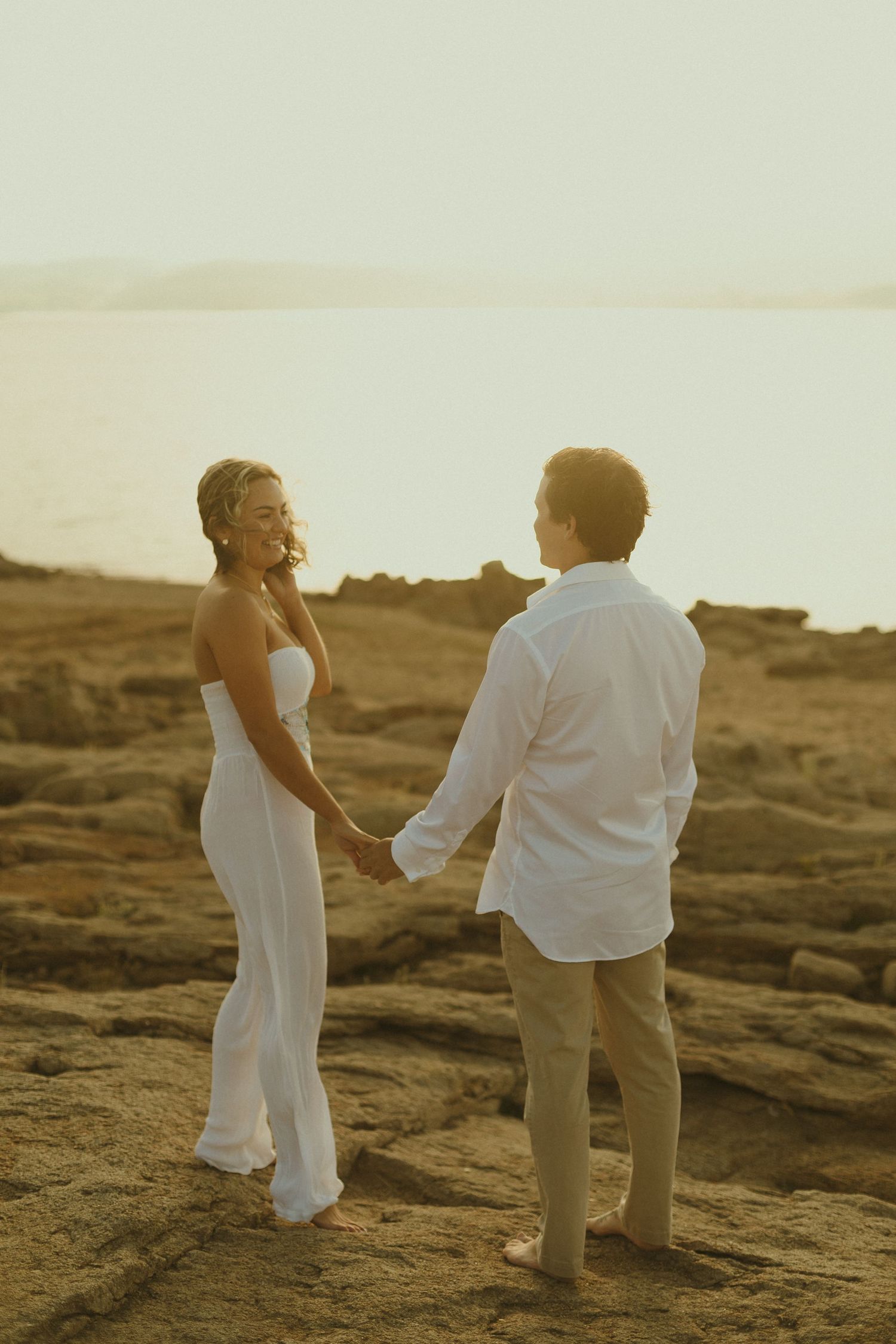 A couple holding hands at sunset on a rocky beach, photographed in warm golden light.
