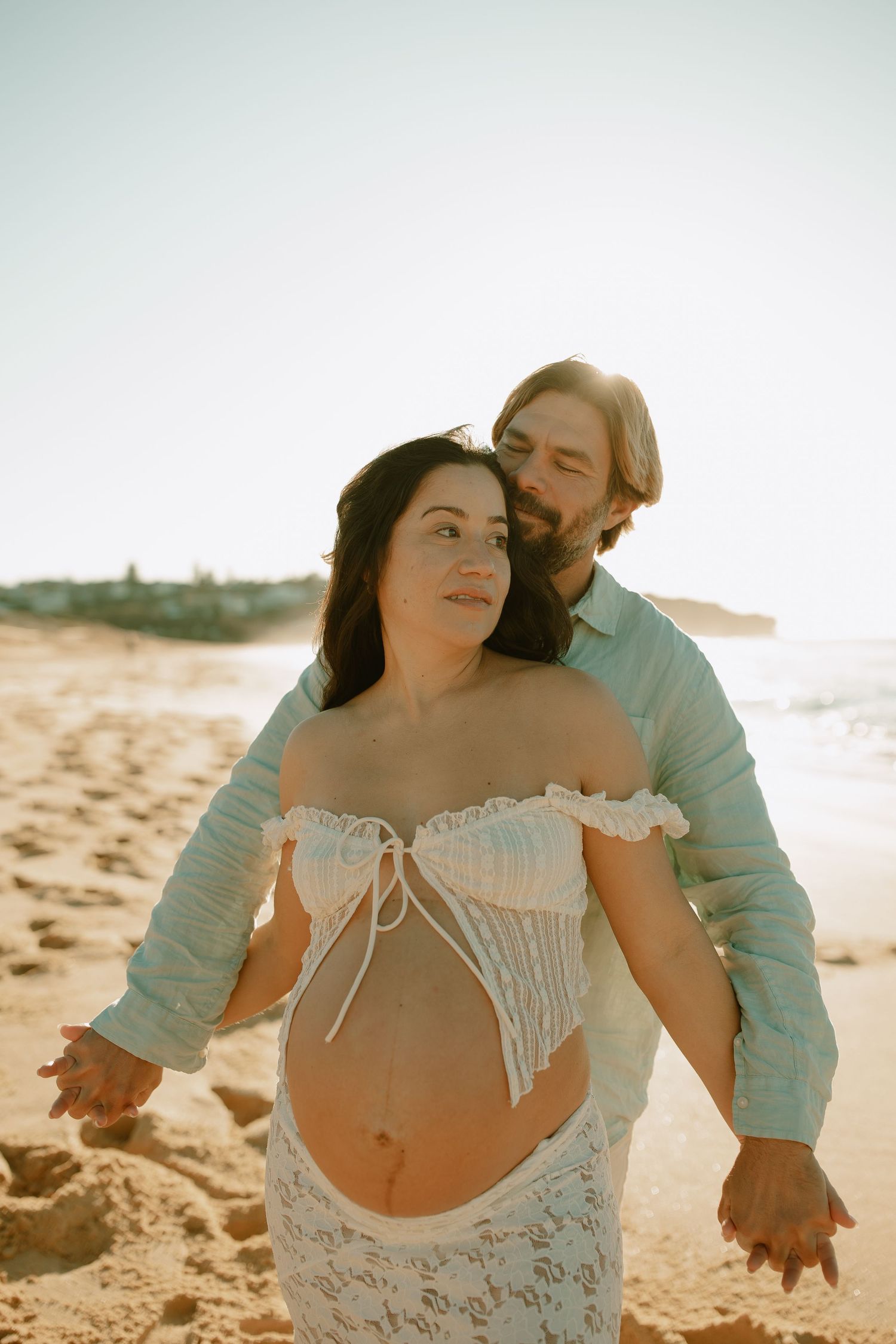 Romantic beach maternity photo with couple in white lace dress and blue shirt.