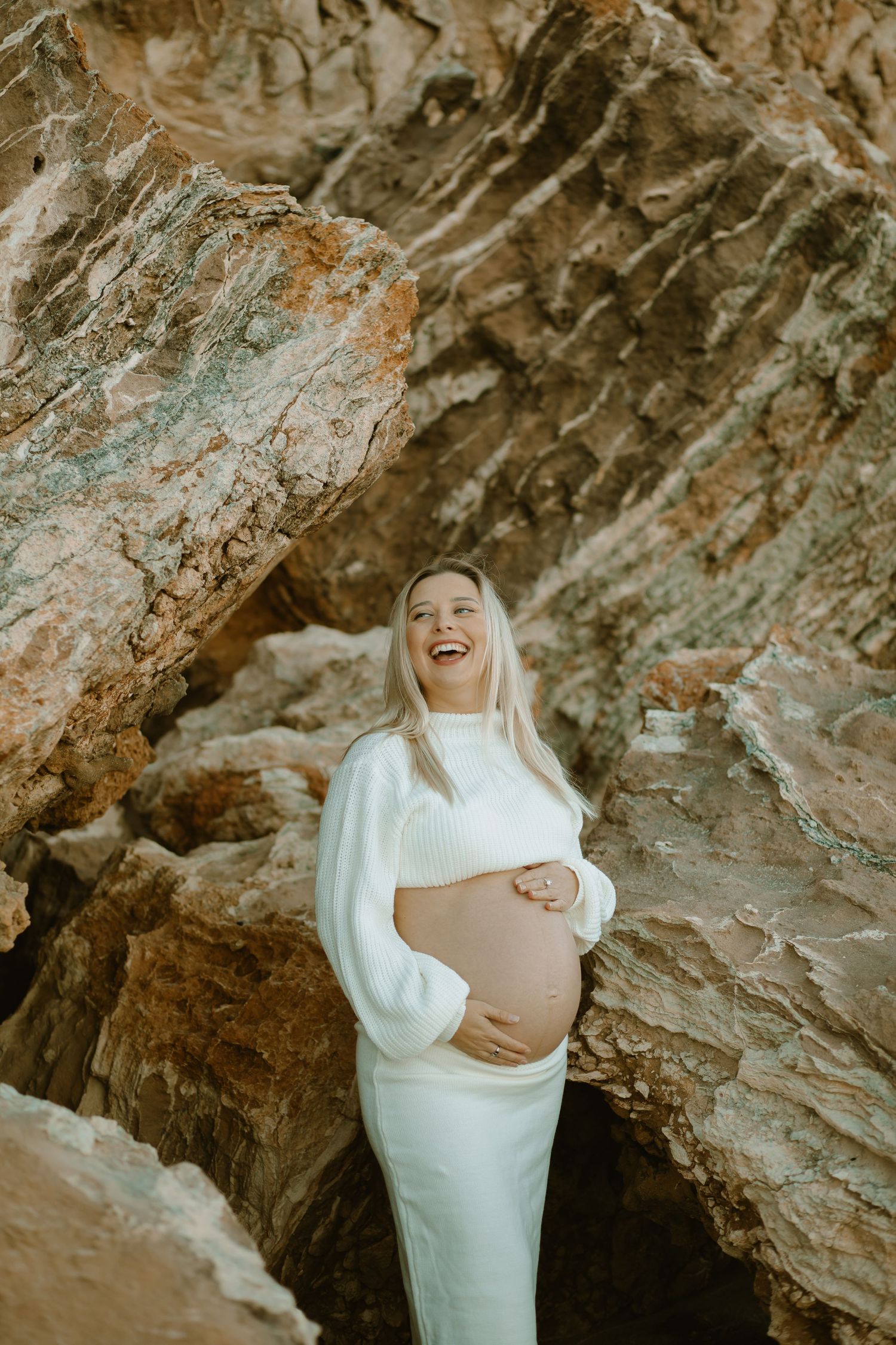 A maternity photoshoot showing a person in a white dress laughing joyfully while standing among dramatic rocky cliffs.
