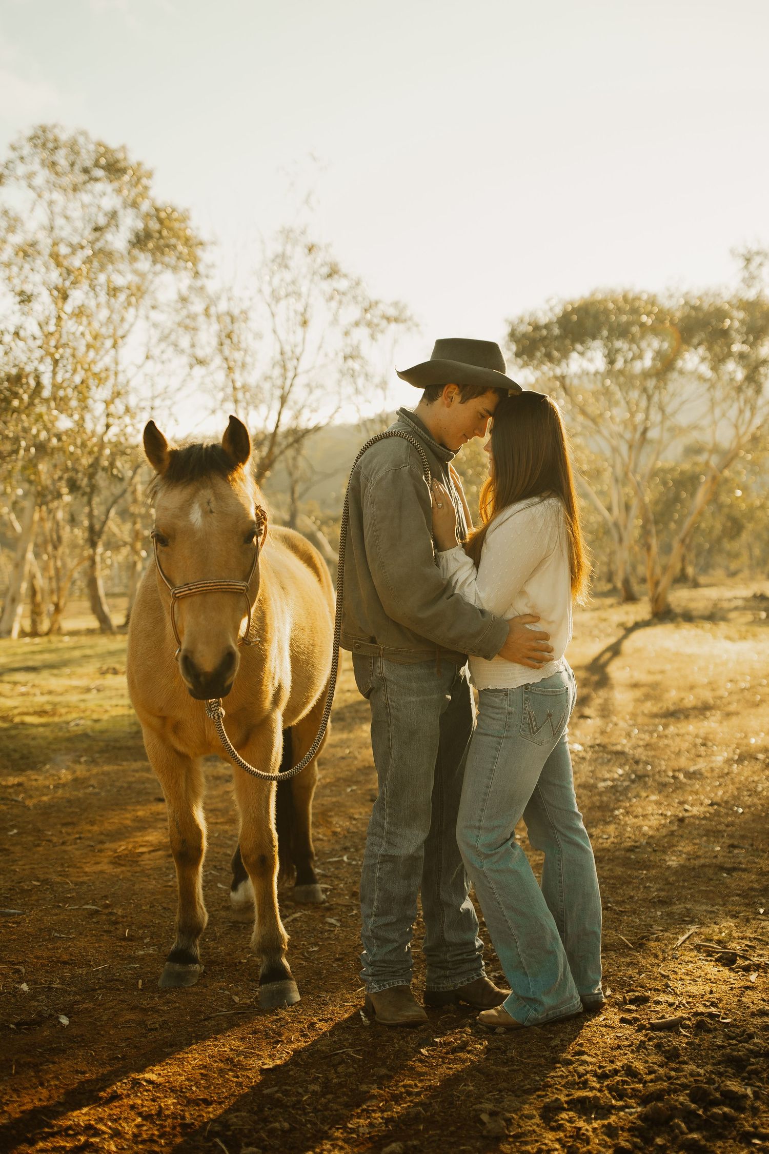 Romantic couple sharing intimate moment next to brown horse on dirt path at sunset in rural setting.
