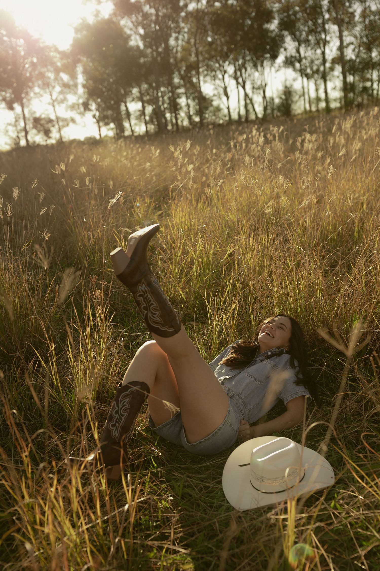 A laughing woman in denim shorts and cowboy boots lies in golden grass field at sunset with white hat beside her.