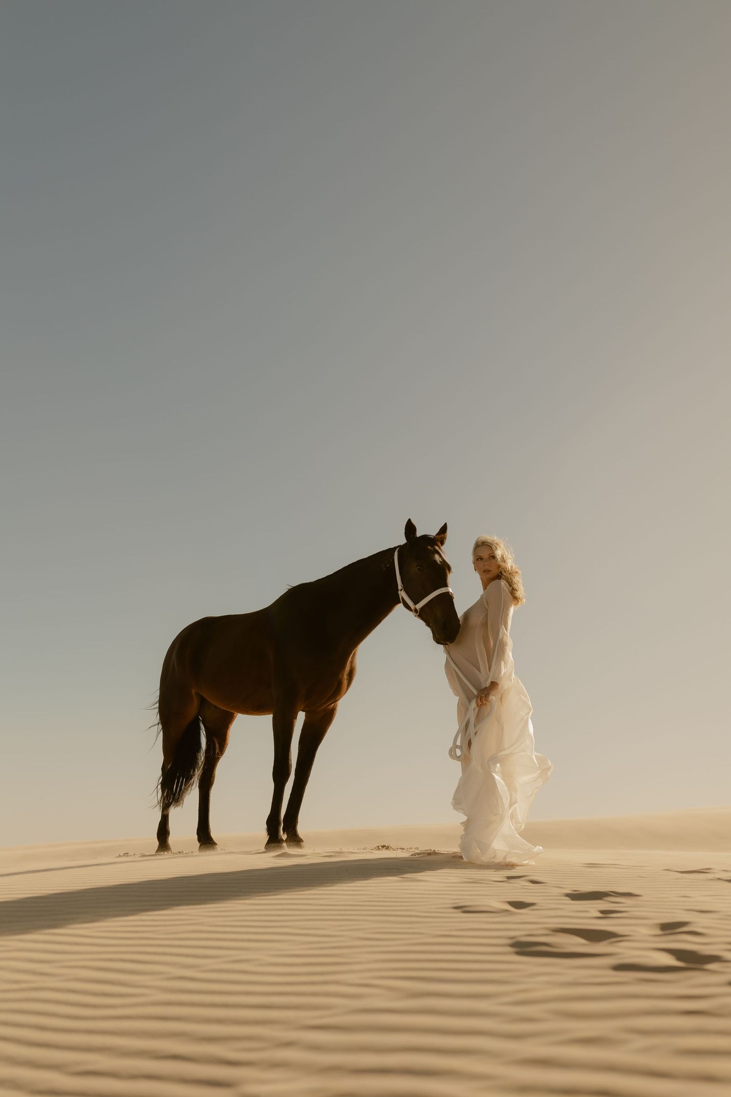 Woman in white dress standing with a dark brown horse on golden sand dunes in a desert landscape.