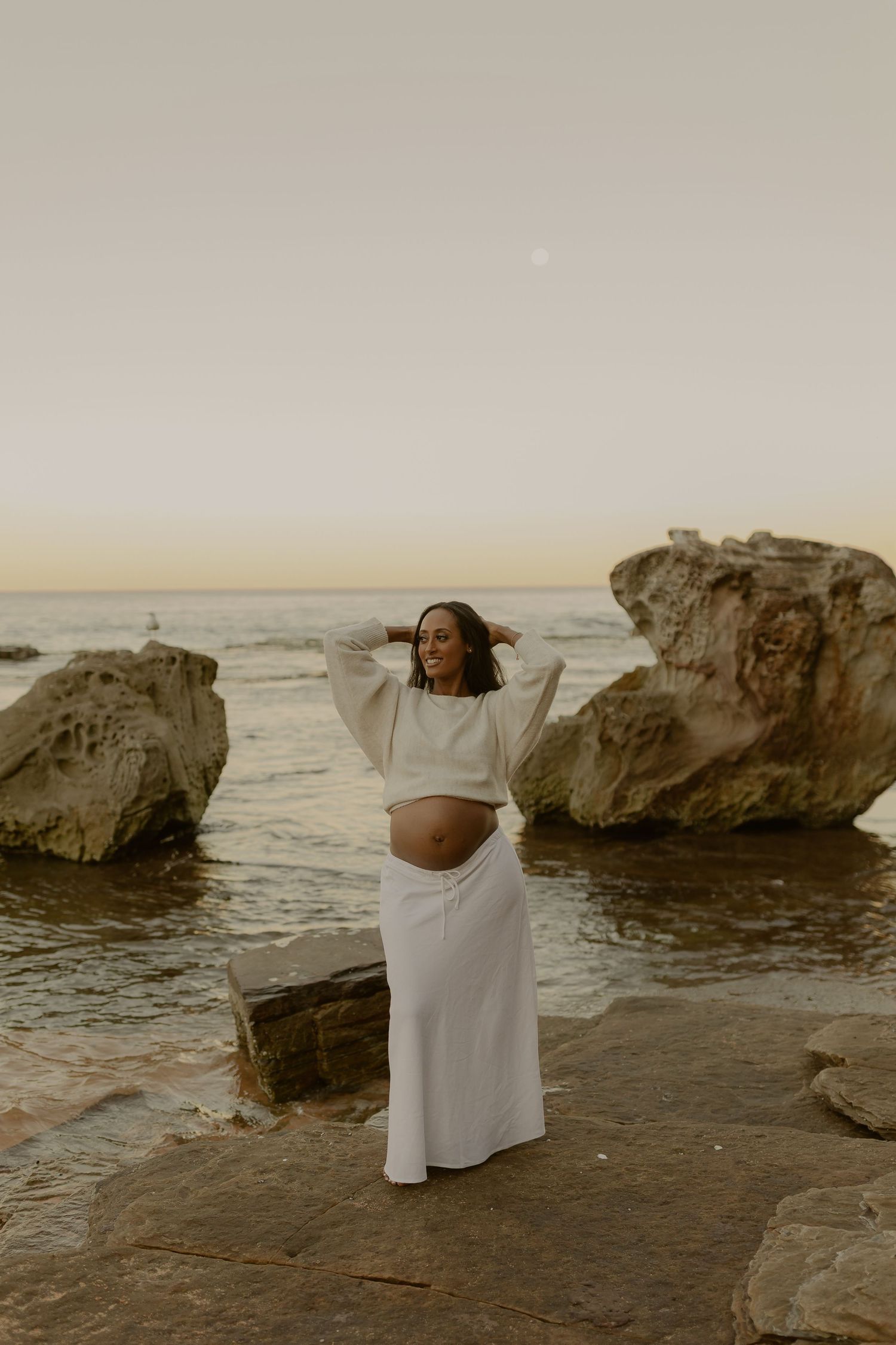 Pregnant woman in white crop top and maxi skirt posing on coastal rocks at sunset.