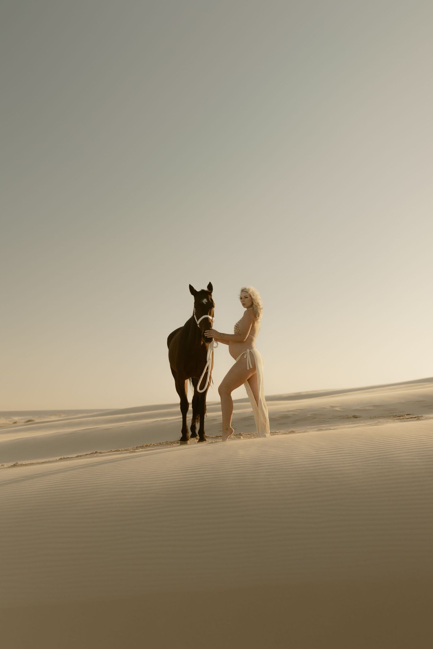 A blonde woman in a white swimsuit stands with a dark horse on sunlit sand dunes under a clear sky.