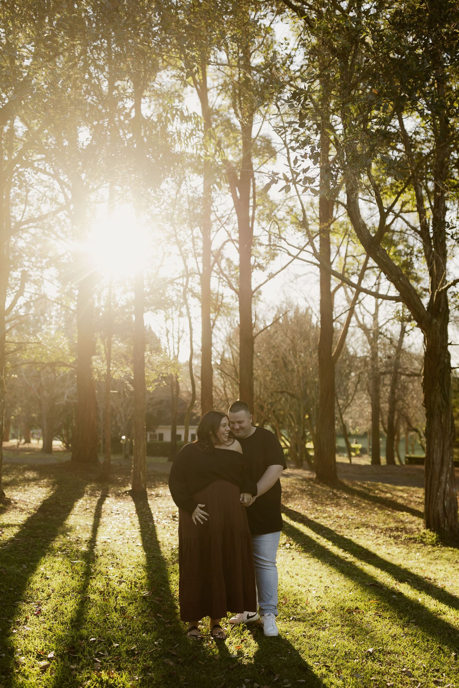 Couple embracing in golden sunlight among tall trees in a park, woman in dark dress with visible baby bump.