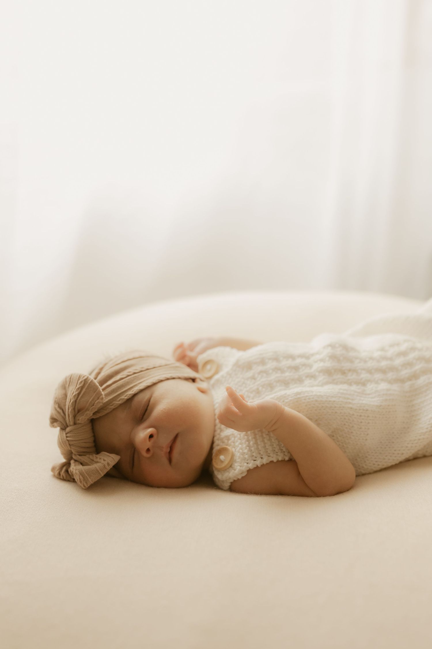 Sleeping newborn baby in white knit romper and beige bow headband on cream surface.