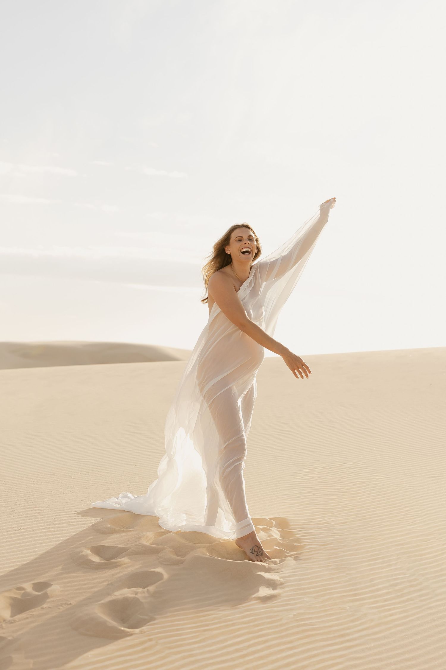 Ethereal figure in flowing white dress dances gracefully across golden sand dunes at sunset.