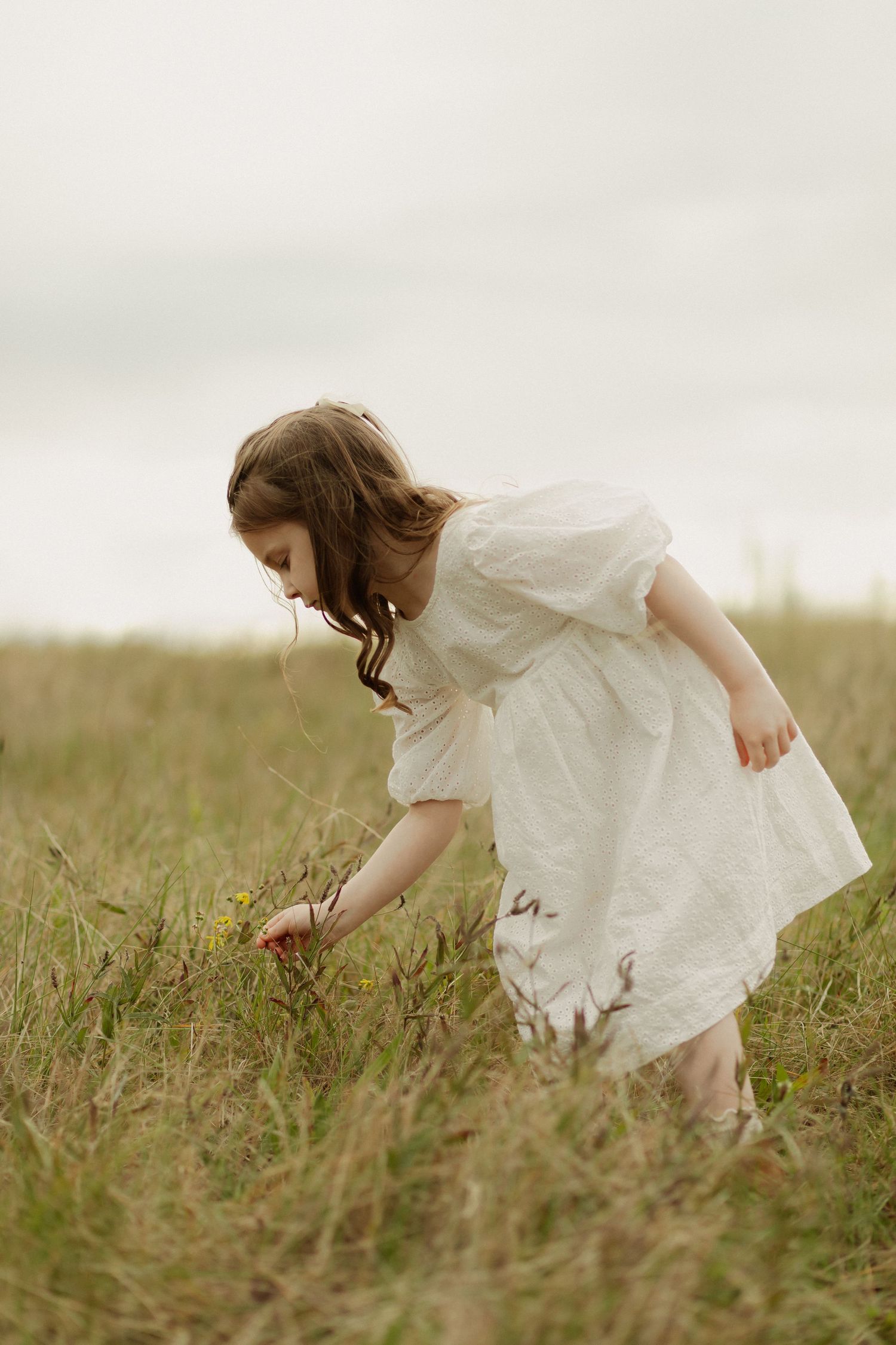 A young girl in a white dress bends down to pick something in a grassy meadow on an overcast day.