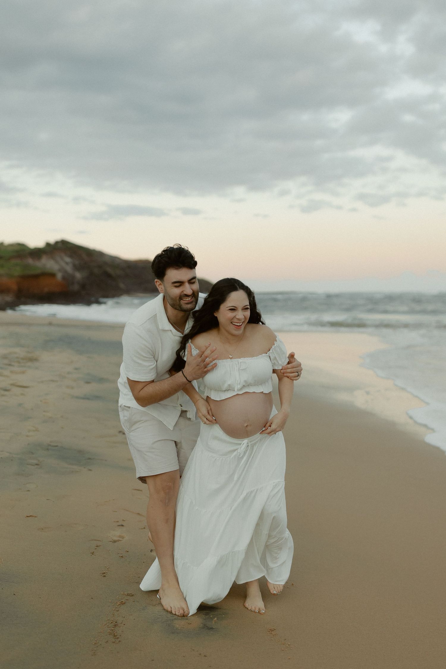 A couple shares intimate moments on a cloudy beach at sunset wearing white flowy outfits.