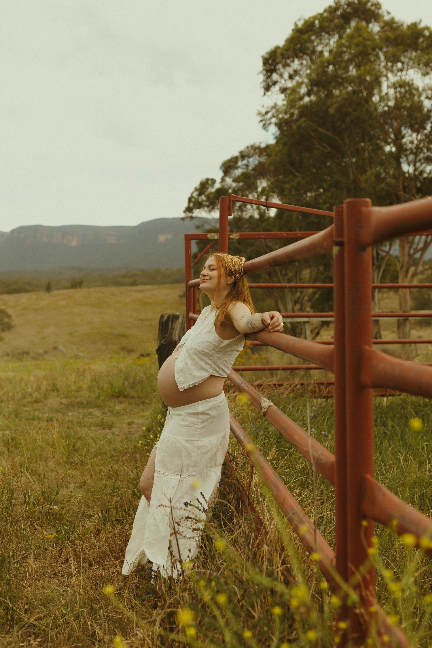 A joyful maternity moment captured against a rustic red gate in a rural landscape.