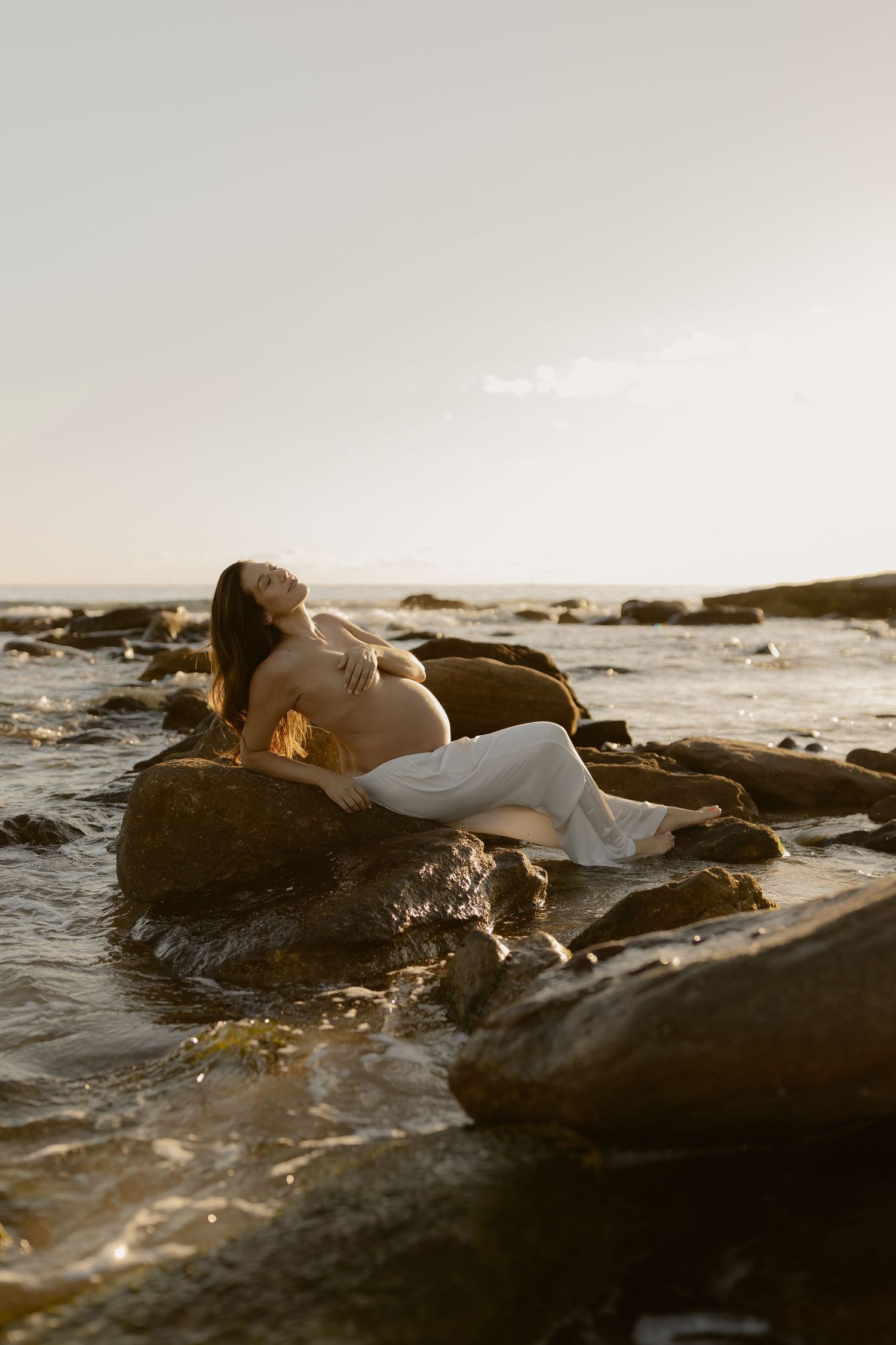 Person in white lying on coastal rocks at golden hour by the ocean.