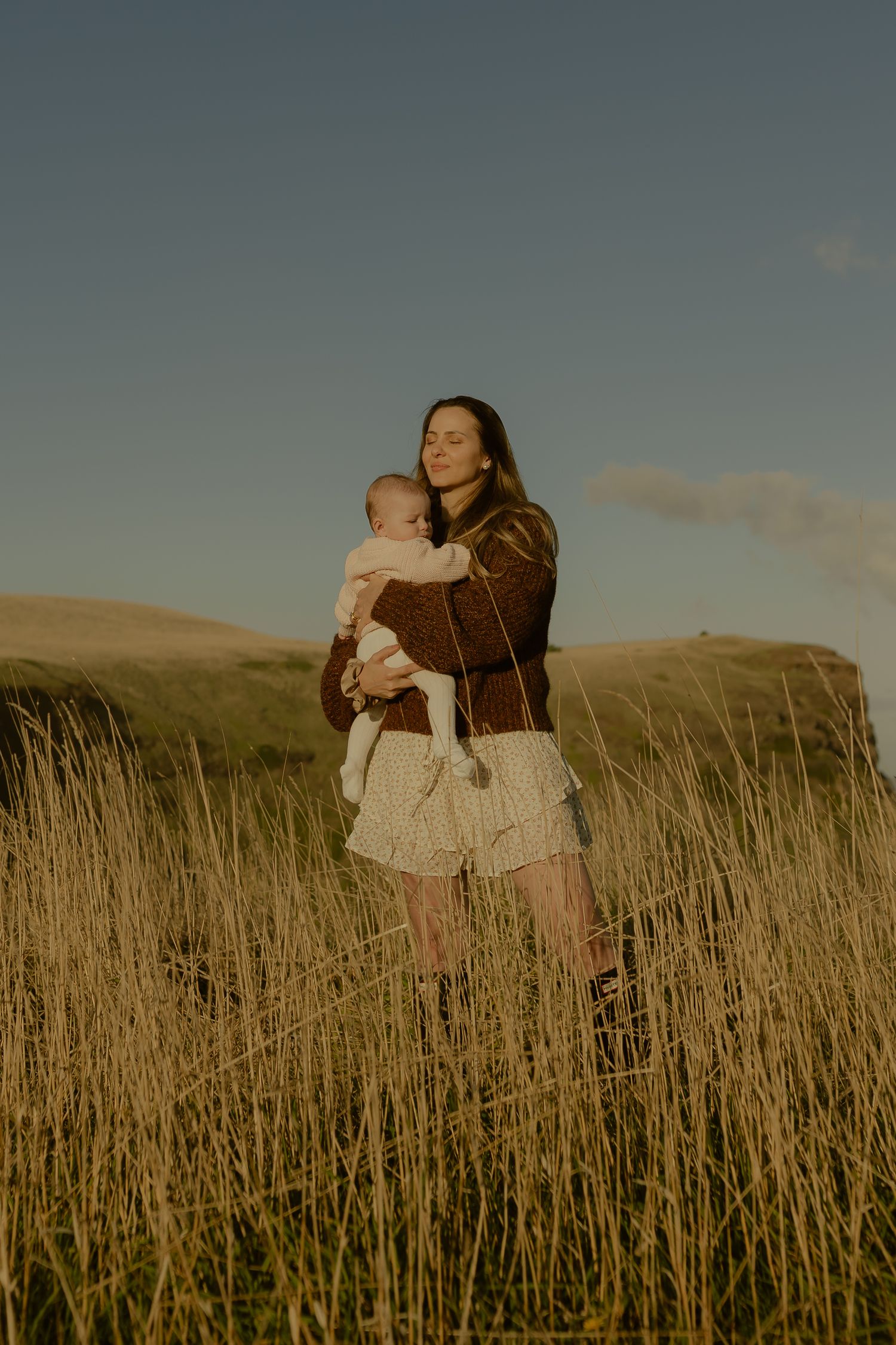 Woman holding newborn baby standing in tall golden grass field at sunset.