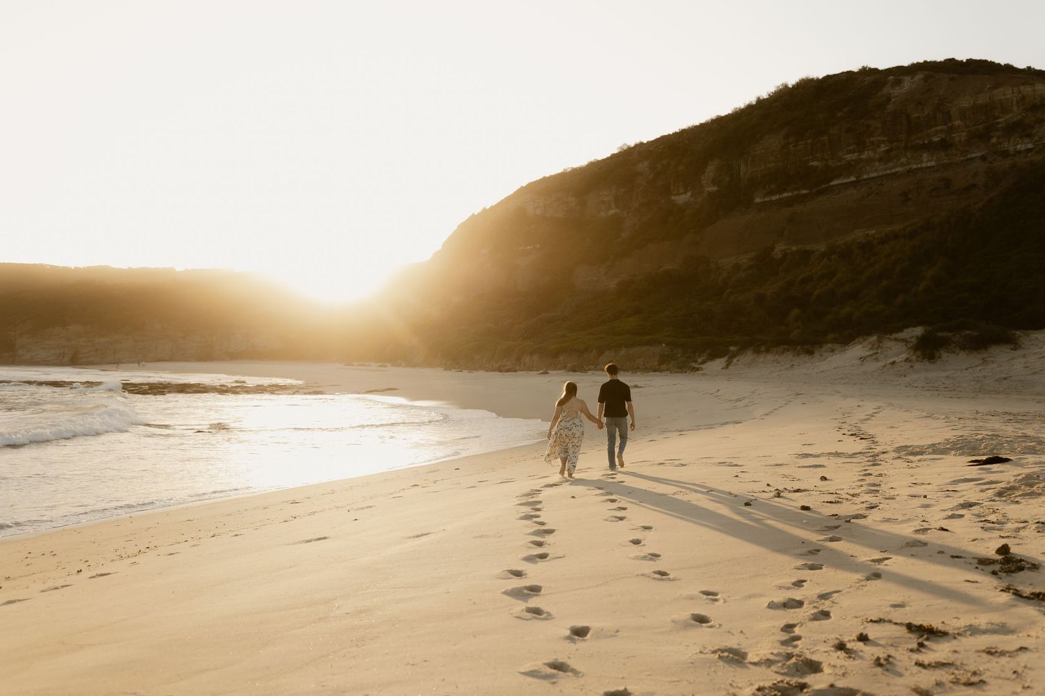 Wide coastal landscape with footprints in the sand leading to distant figures on the beach.