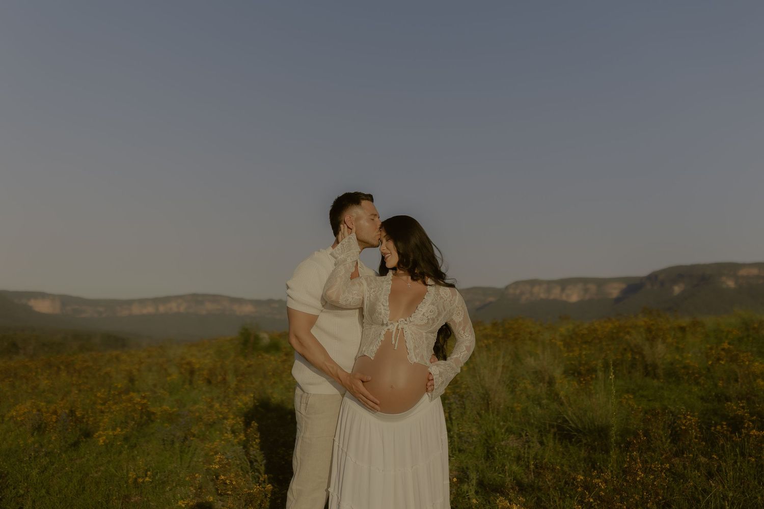 A tender moment captured between a couple in white attire against a mountainous landscape at dusk.