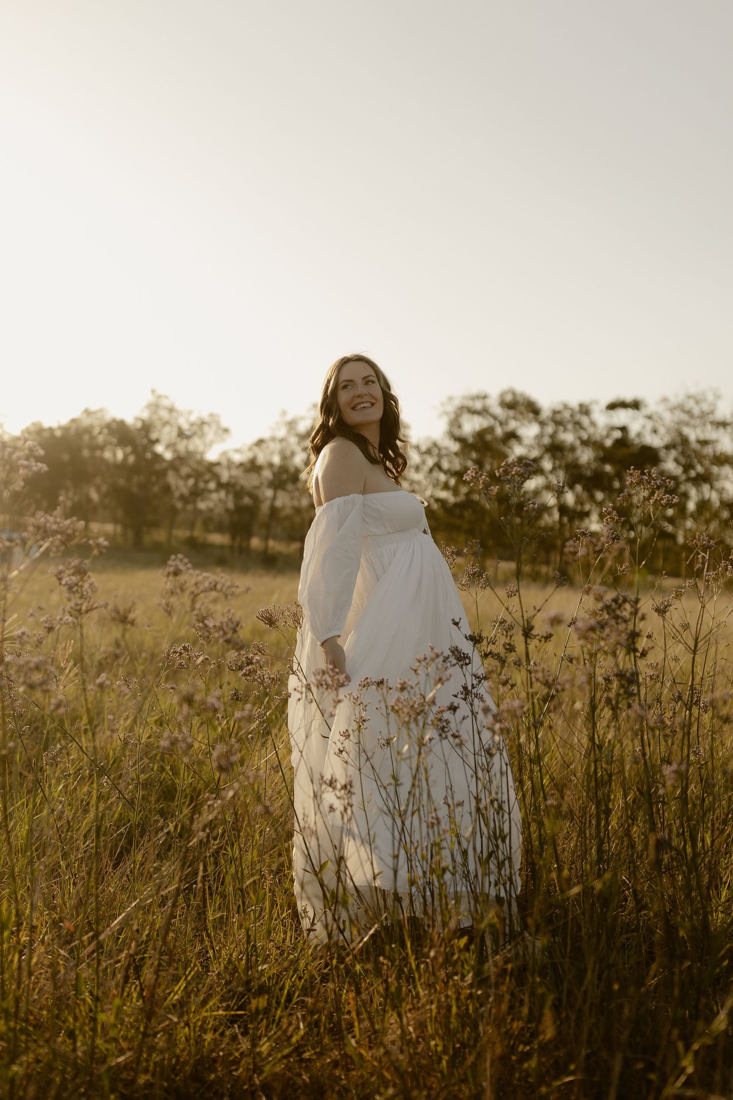 A figure in a flowing white dress stands in a golden meadow at sunset among tall wildflowers and grasses.