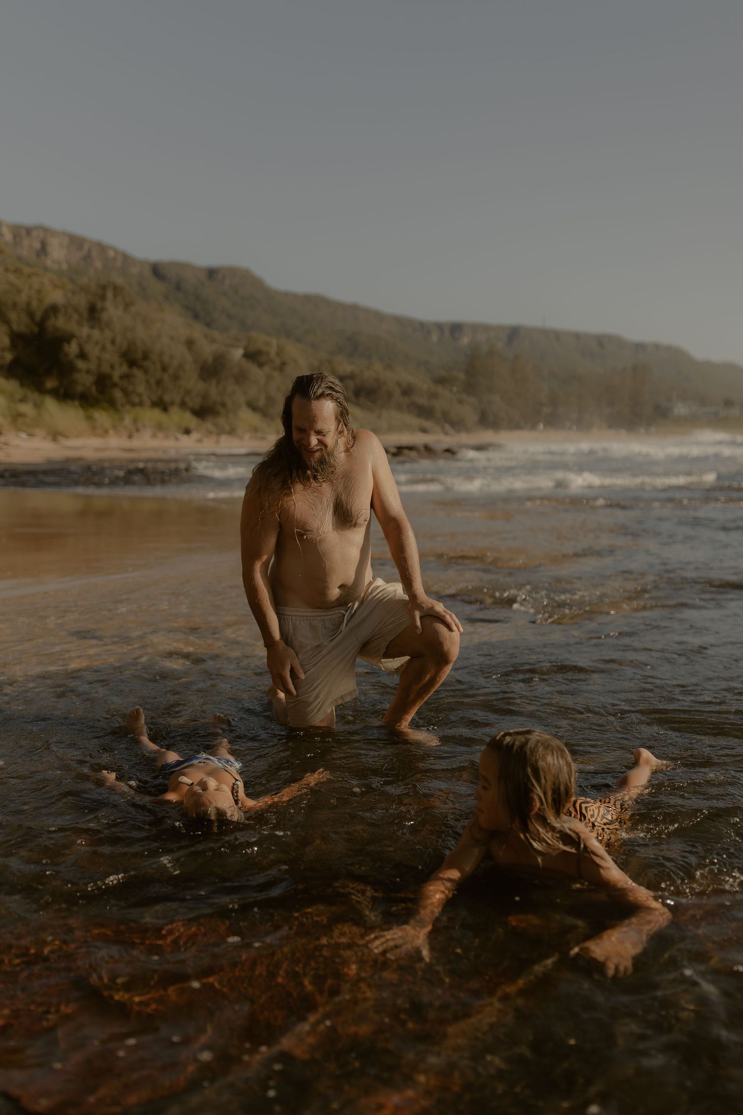 Friends playing and splashing in shallow ocean water near a rocky coastline at dusk.
