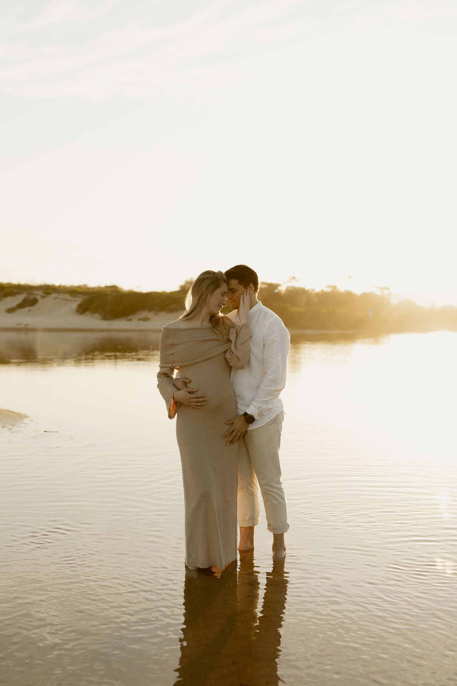 A couple embraces while standing in shallow water at sunset by the shore.