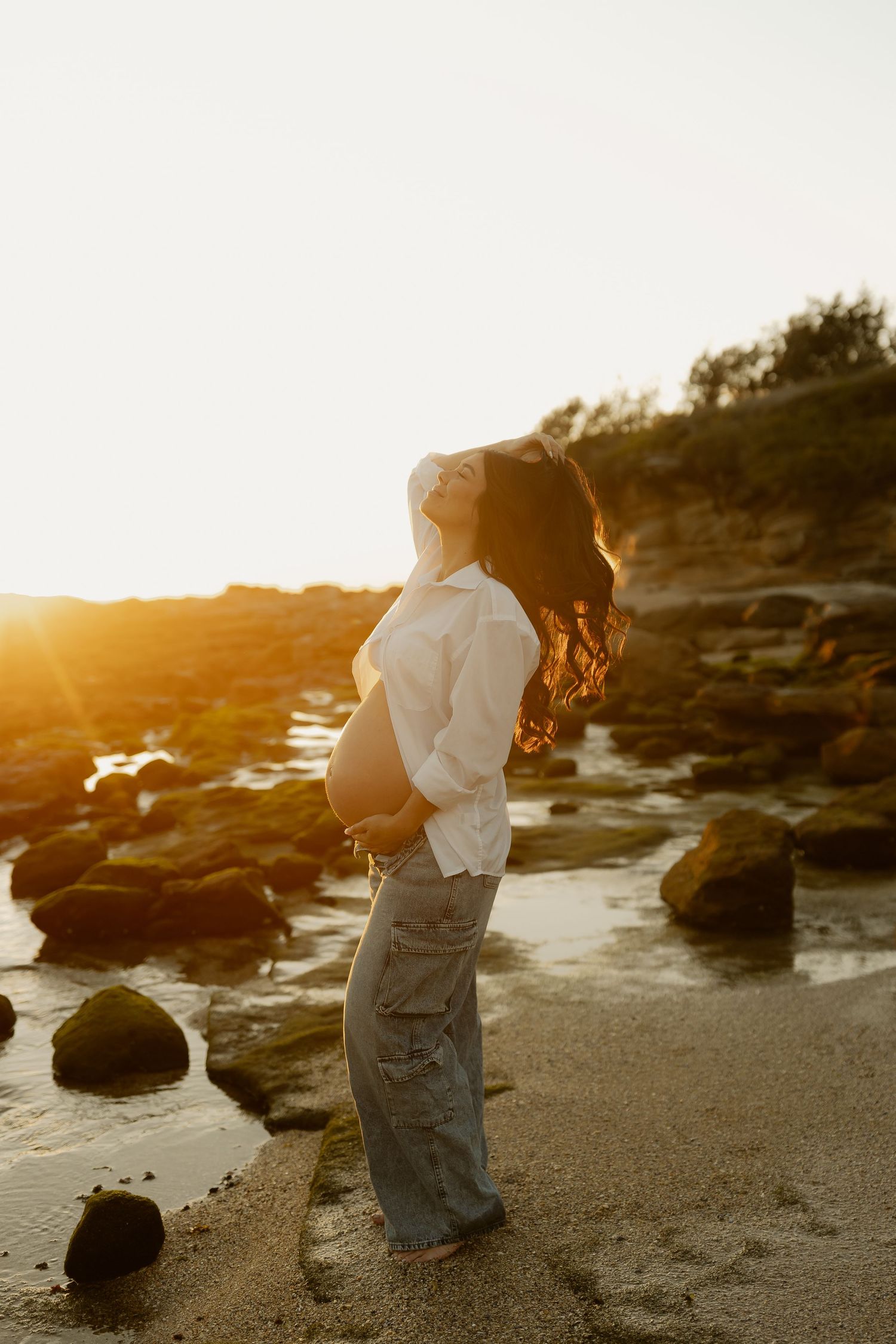 Pregnant woman in white shirt and jeans posing on rocky beach during stunning golden sunset.