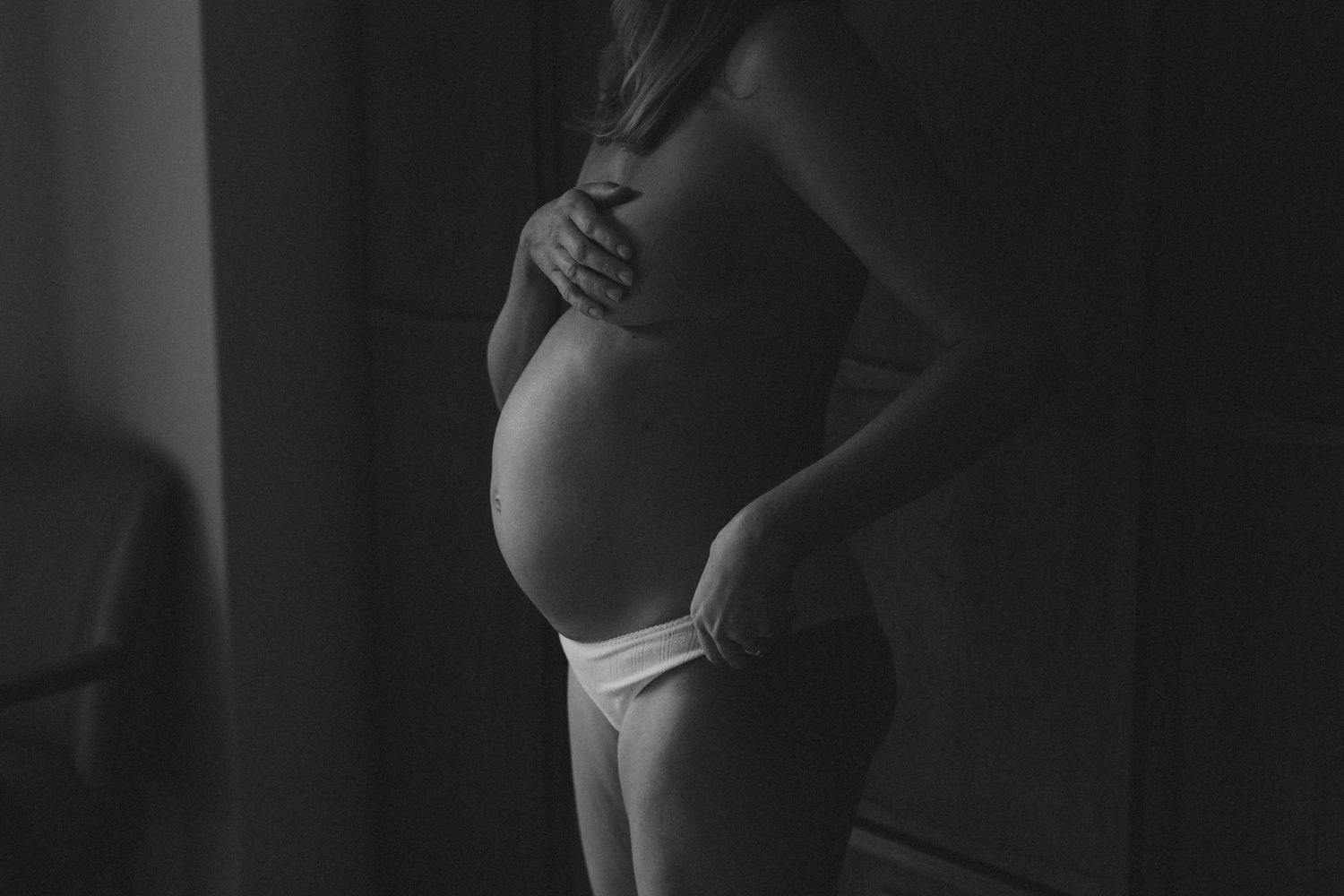 Black and white maternity photo of pregnant woman cradling bare baby bump in underwear.