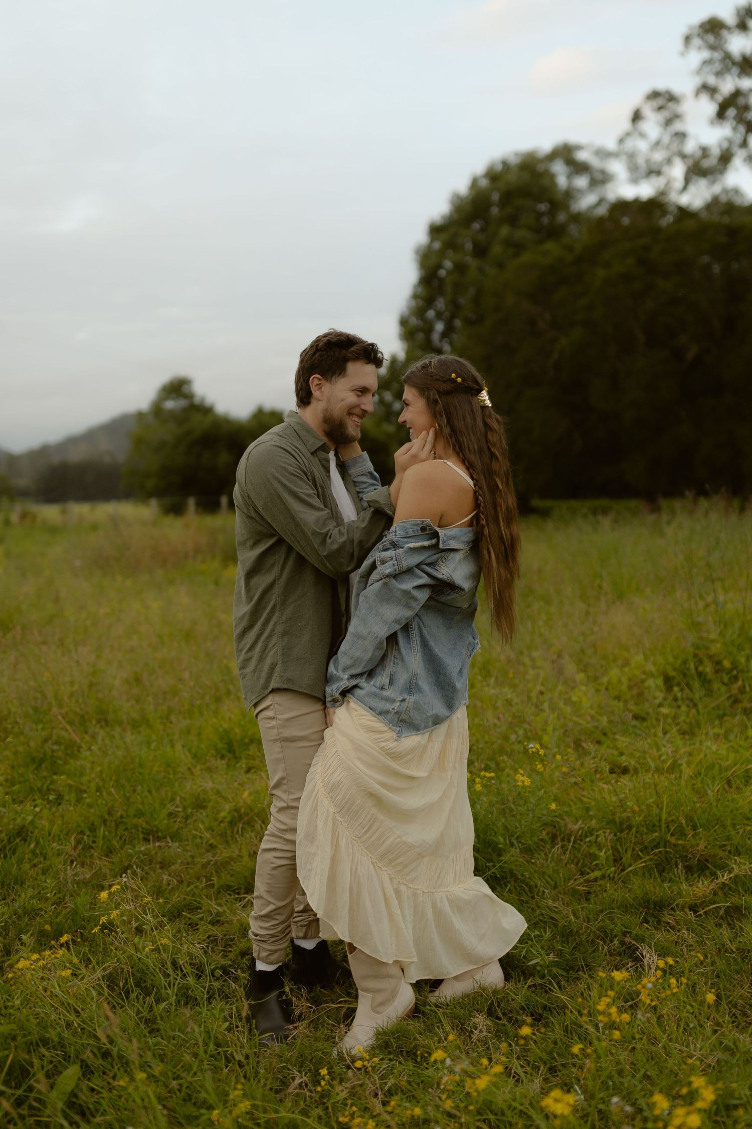 Romantic couple embracing and smiling in a lush green meadow, woman wearing off-shoulder denim jacket and cream dress.