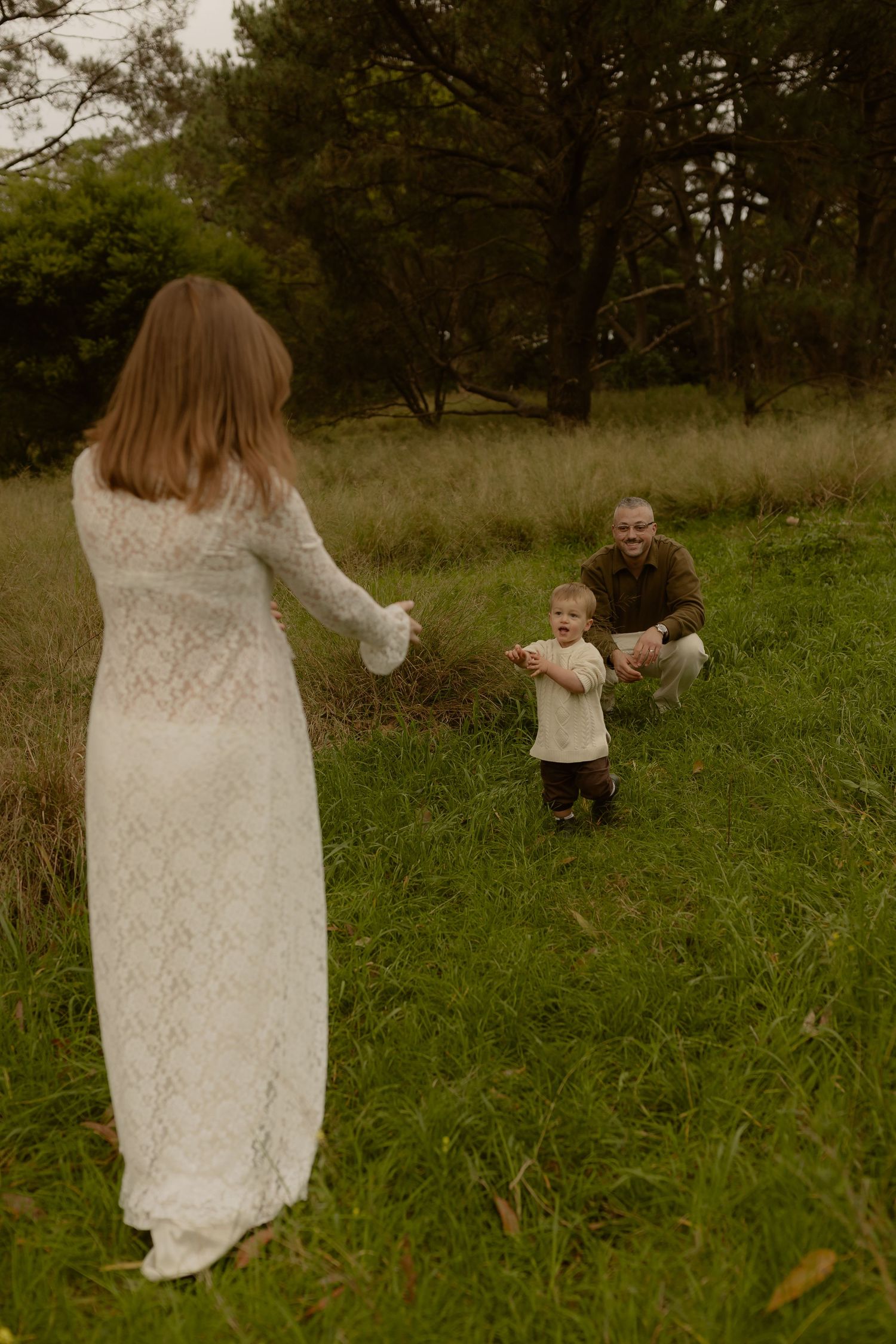 A family maternity photo session outdoors, with a toddler walking toward their pregnant mother in a white lace dress.