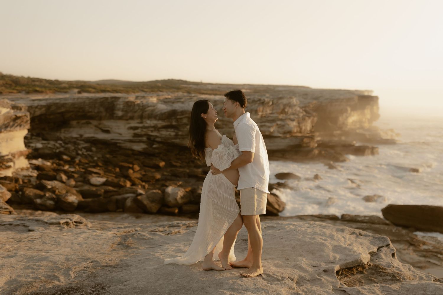 Romantic couple embracing on rugged coastal cliffs during a golden sunset.