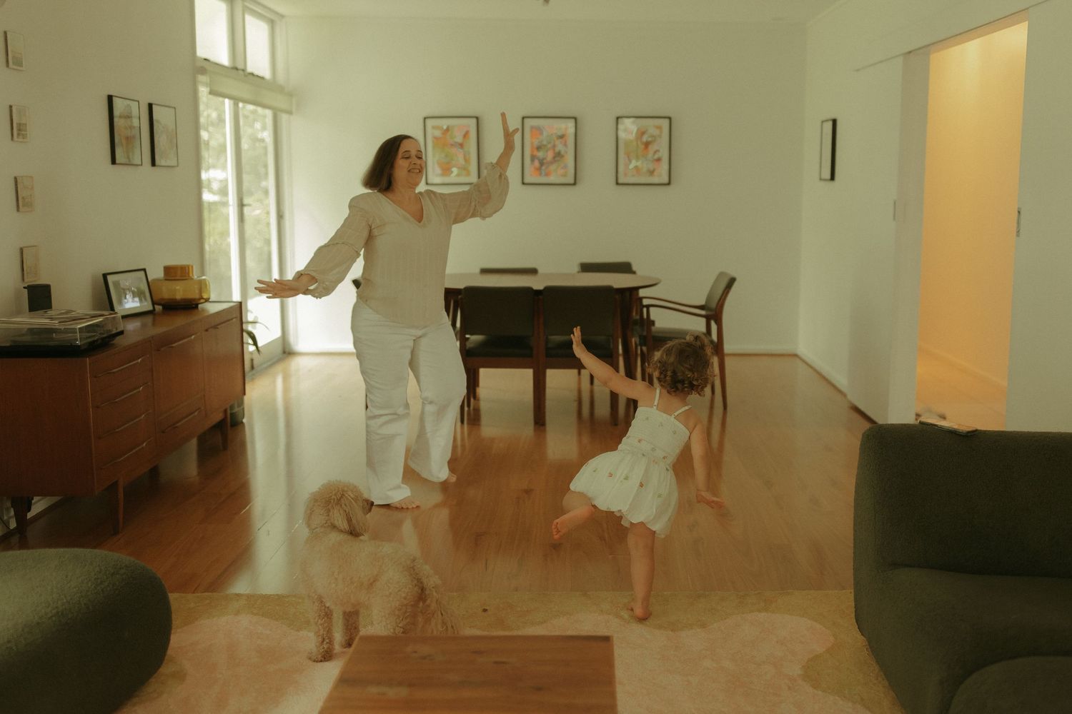 Two people dance together in a bright living room with hardwood floors and a piano in the background.