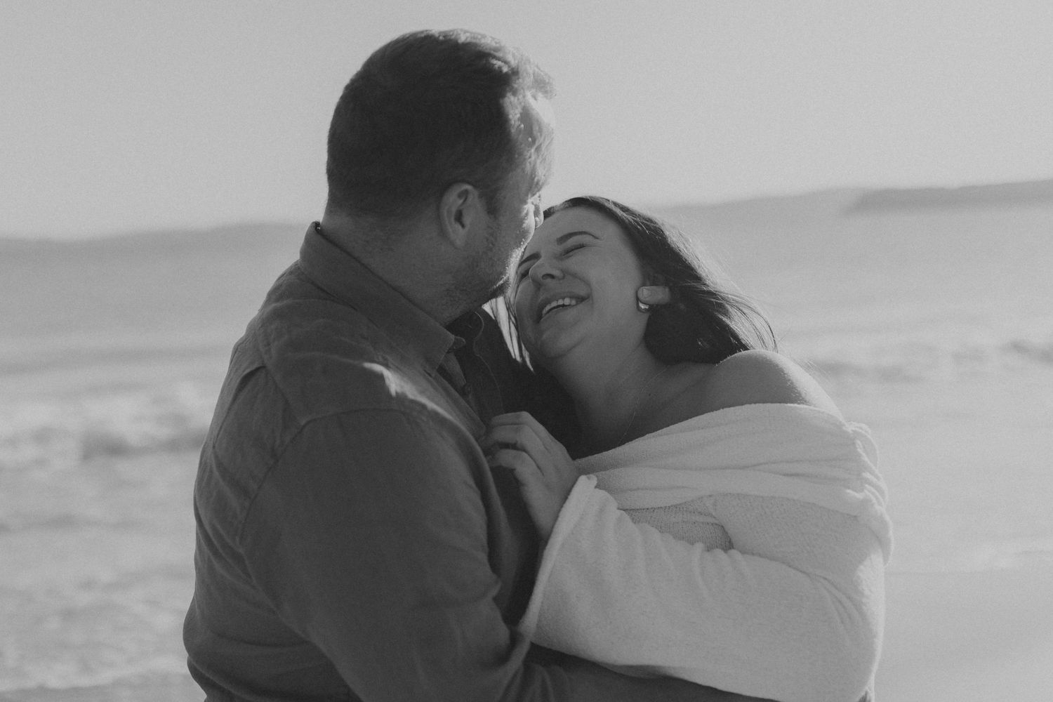 A couple shares a joyful moment together while embracing on a beach in black and white.