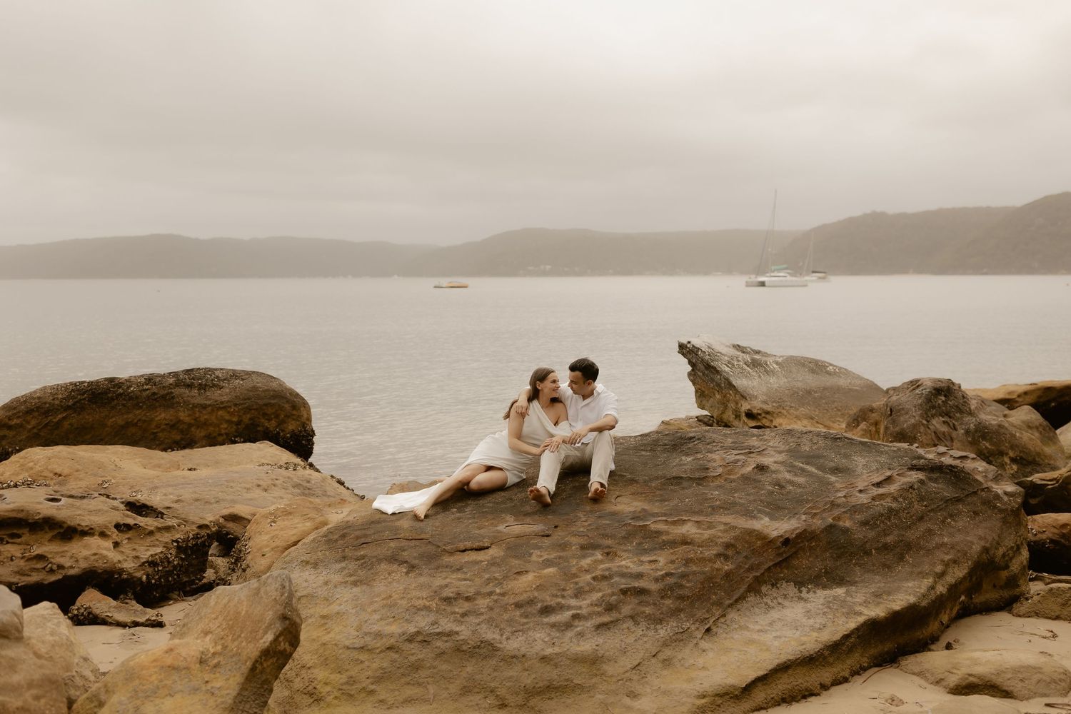 A couple sits together on large coastal rocks overlooking a misty bay and distant mountains.